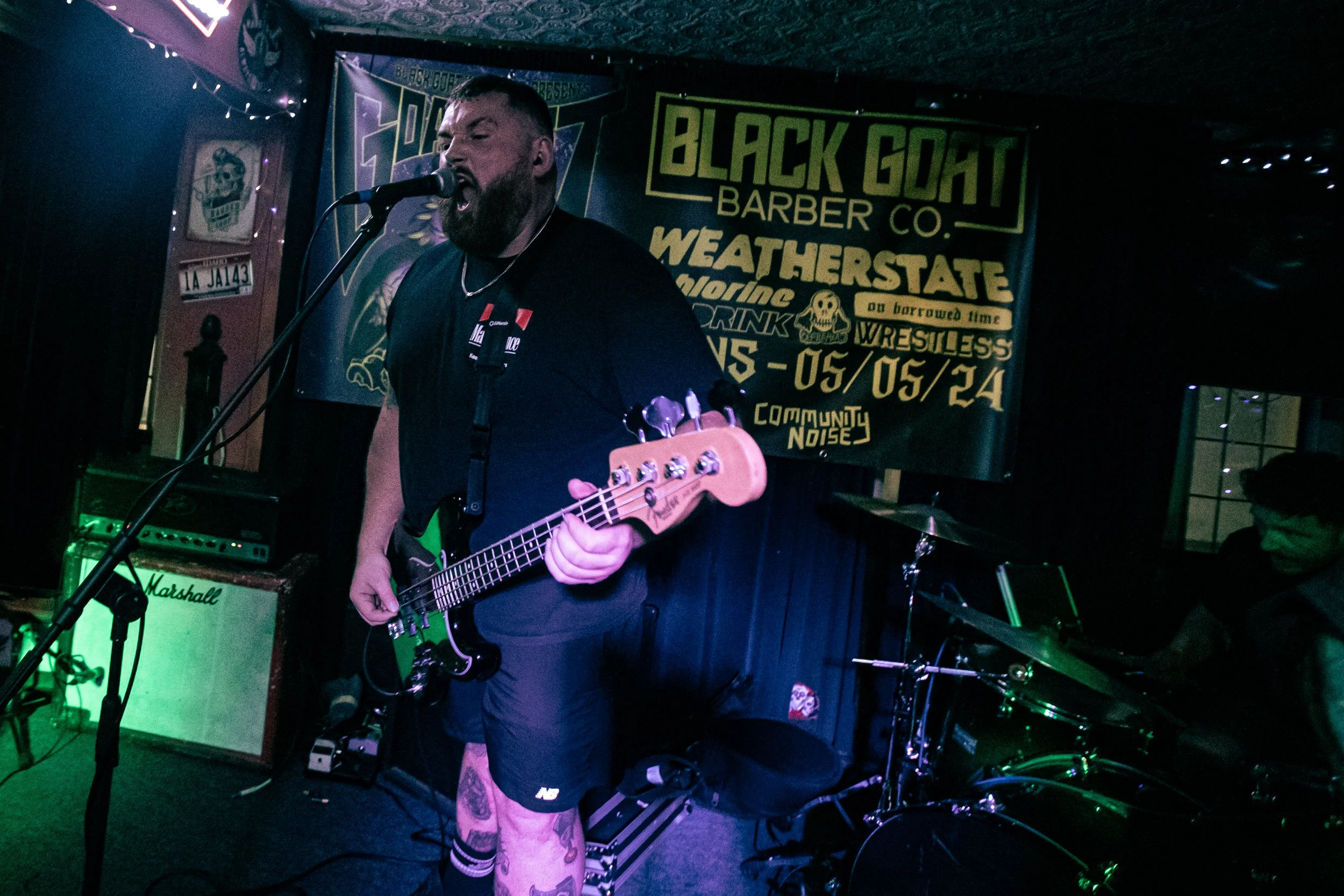 A man singing into a microphone while playing an electric bass guitar on stage with a drummer in the background at a live music event.