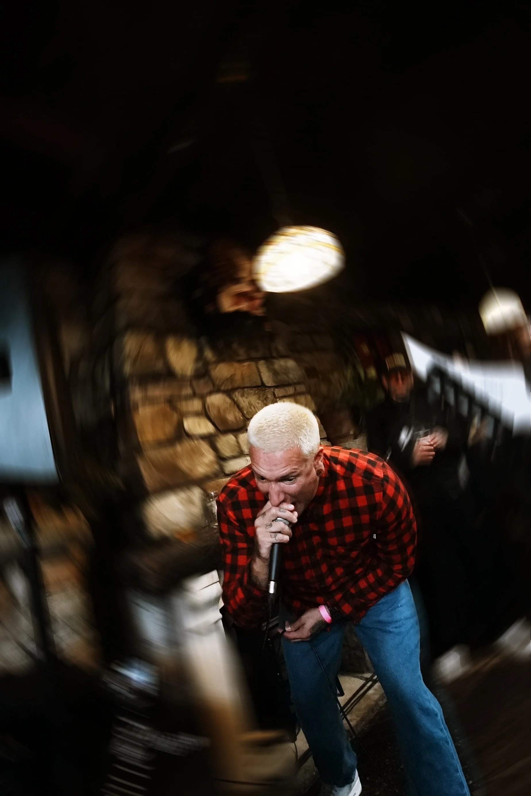 A person with platinum blonde hair singing into a microphone in a dimly lit venue with a stone wall background.