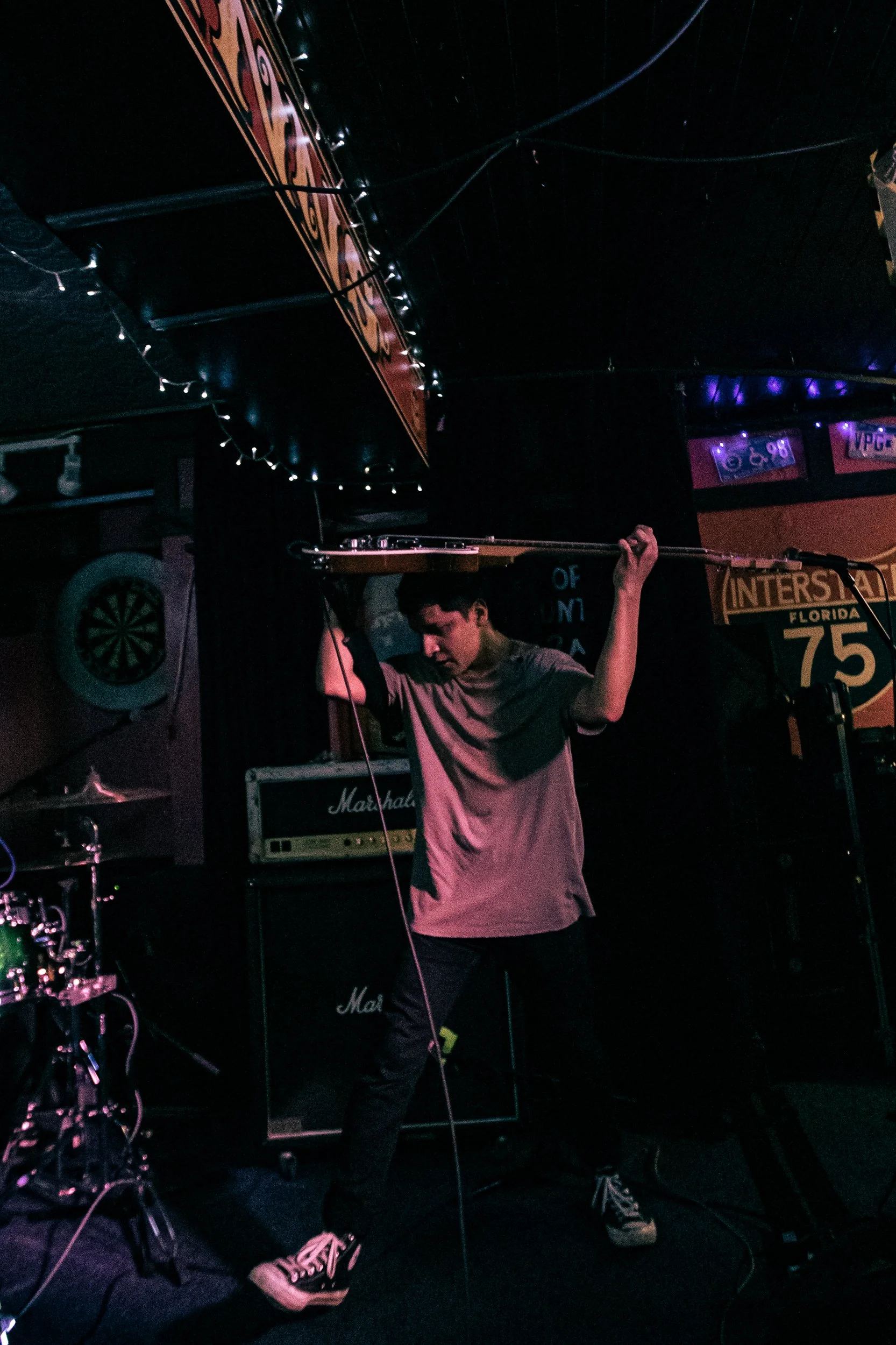 A young man holding a guitar in a darkened stage area, with Marshall amplifiers, a dartboard, and neon lights in the background.
