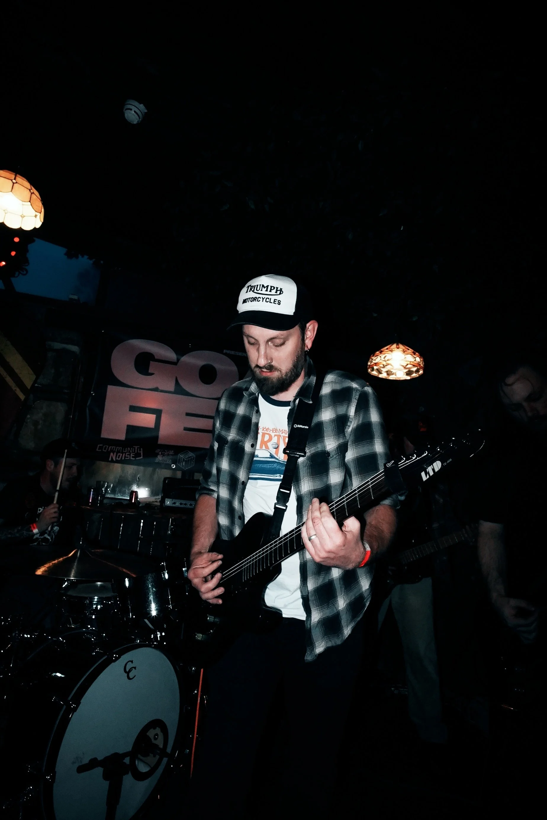 Musician playing electric guitar at a dimly lit venue with a sign reading 'GONE FEST' in the background.