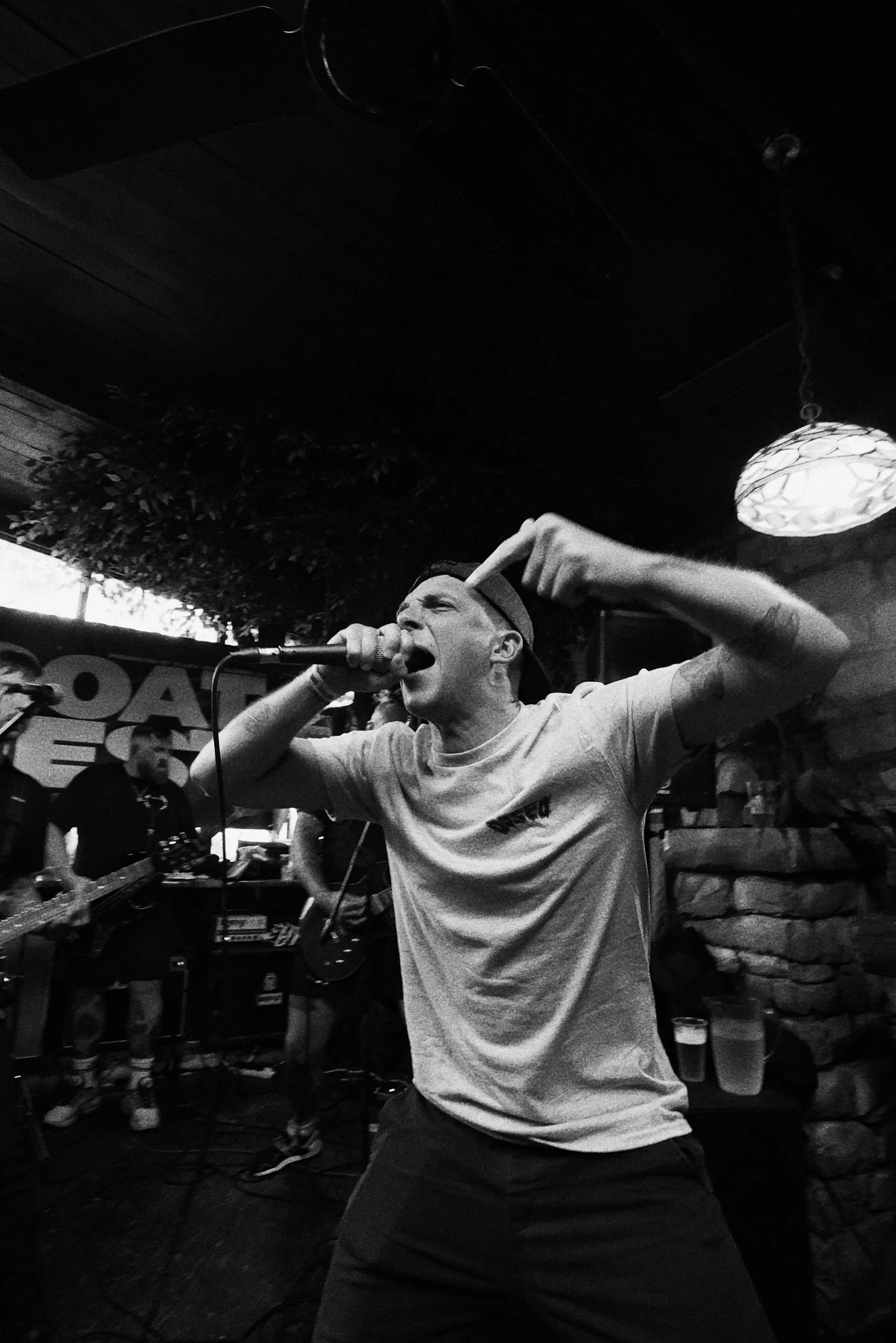 A man passionately singing into a microphone during a live music performance in a dimly lit venue, with band members and musical equipment in the background.