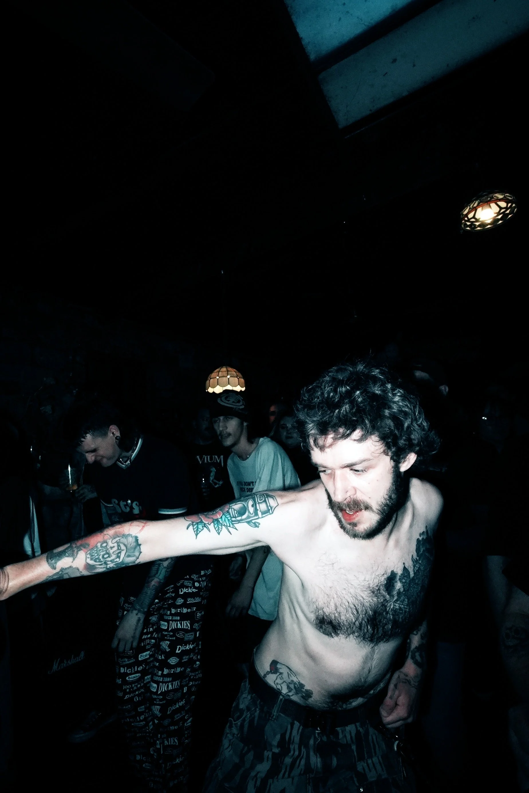 A shirtless man with curly hair and tattoos extending from his arm to his chest and stomach dancing in a dark, crowded room with low lighting and vintage-style ceiling lights.