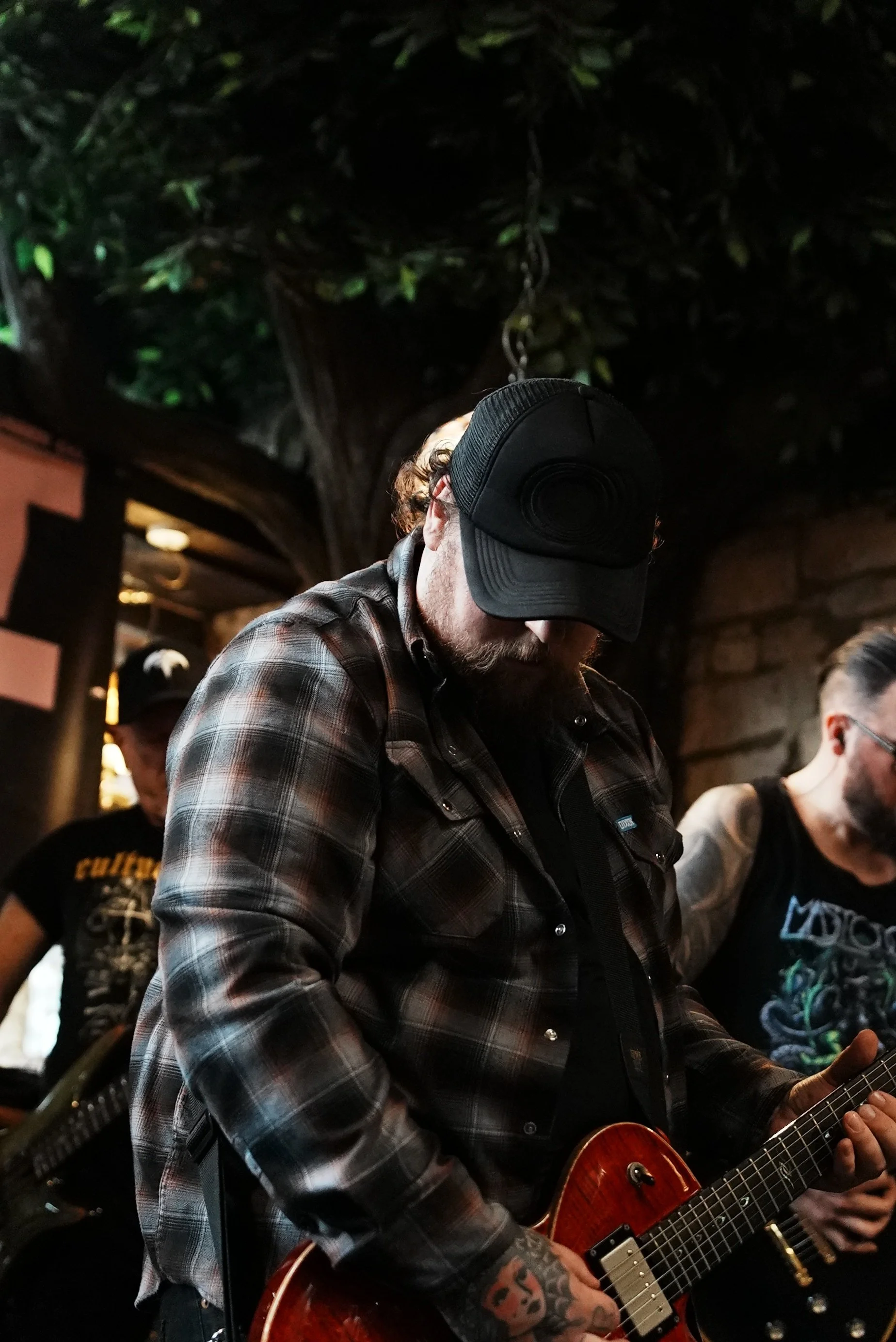 A man playing an electric guitar with a tattoo on his hand, wearing a plaid shirt and cap, in a dimly lit indoor setting.