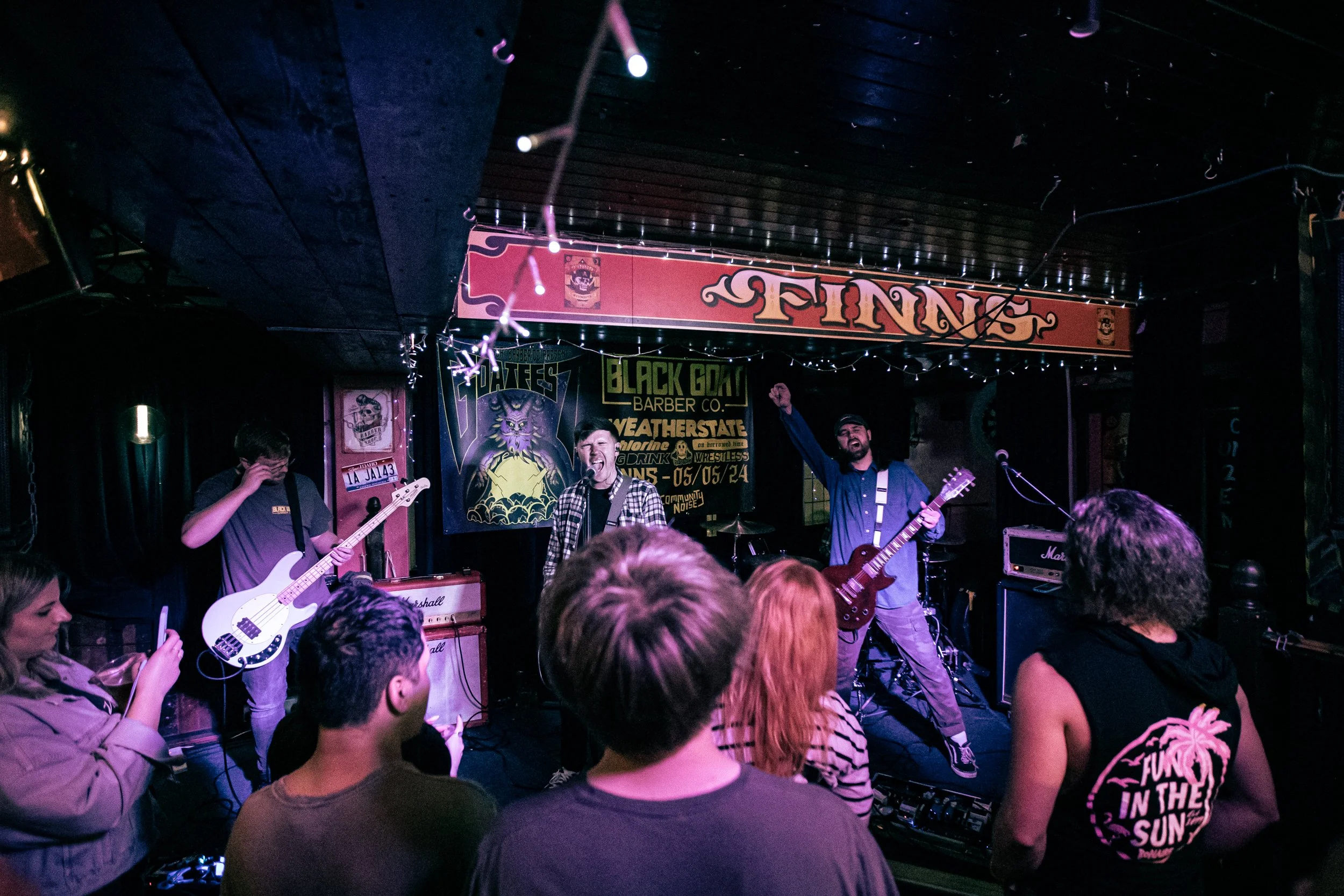 A band performs on stage at a bar or club, with three band members playing guitars and singing into microphones. Audience members are watching, one person is taking a photo, and the scene is lit with colorful stage lighting and string lights.
