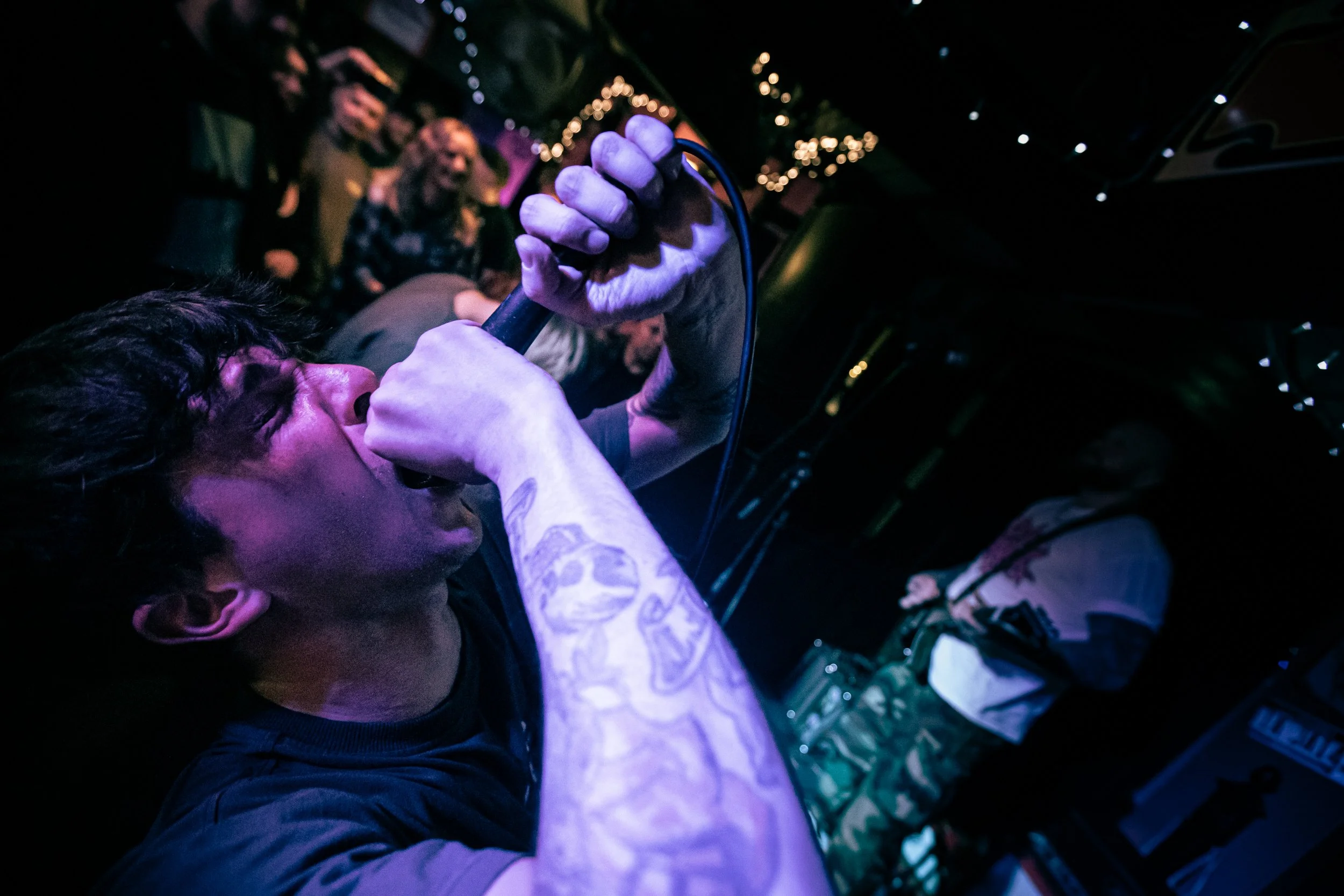 A male singer with tattoos singing into a microphone at a music venue, with a small audience watching.