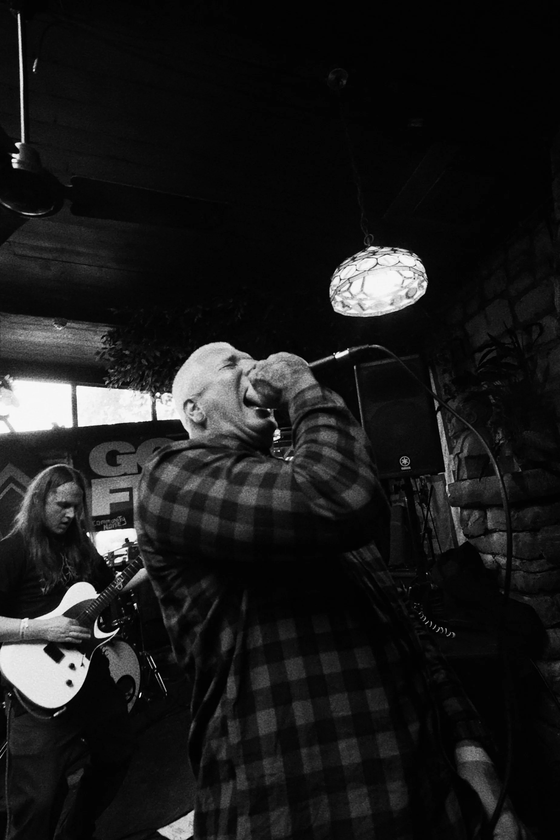 A man with short hair singing into a microphone and a guitarist with long hair playing an electric guitar in a dimly lit venue.