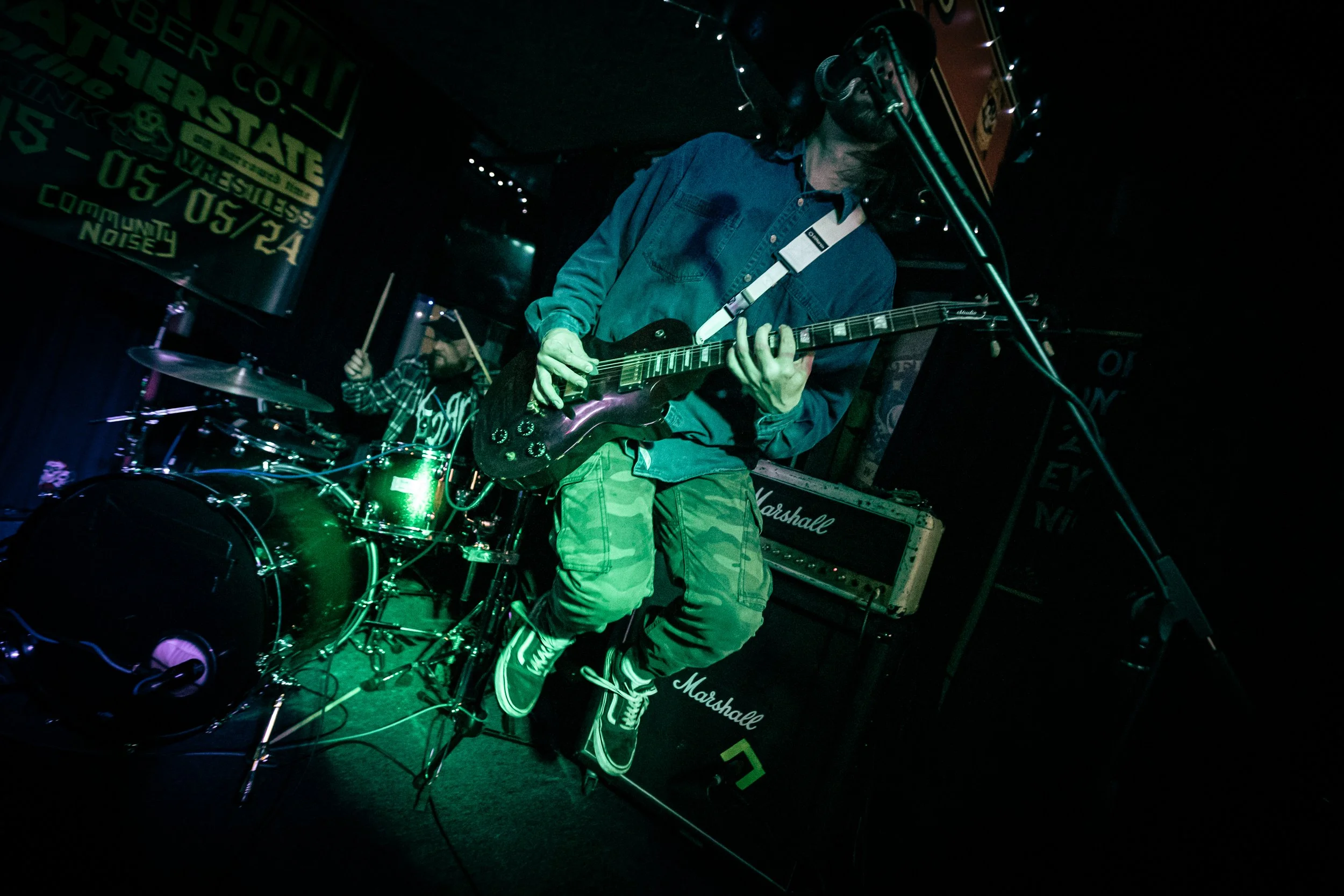Musicians performing on stage with a guitarist in the foreground and a drummer in the background, stage lit with colored lights, and Marshall amplifiers visible.