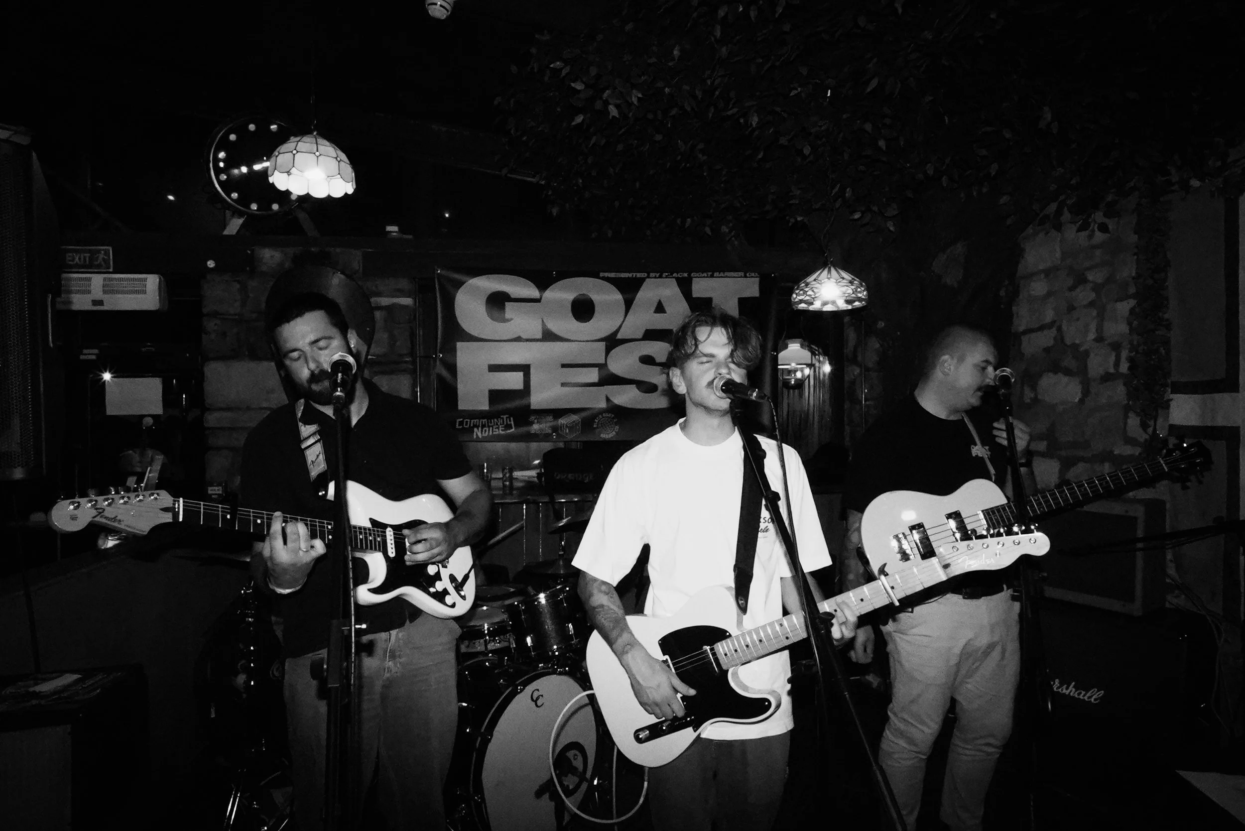 A band performing live on stage at a music venue, with three members playing guitars and singing into microphones. A banner in the background reads 'GOAT FEST' and the venue has stone walls and hanging lamps.