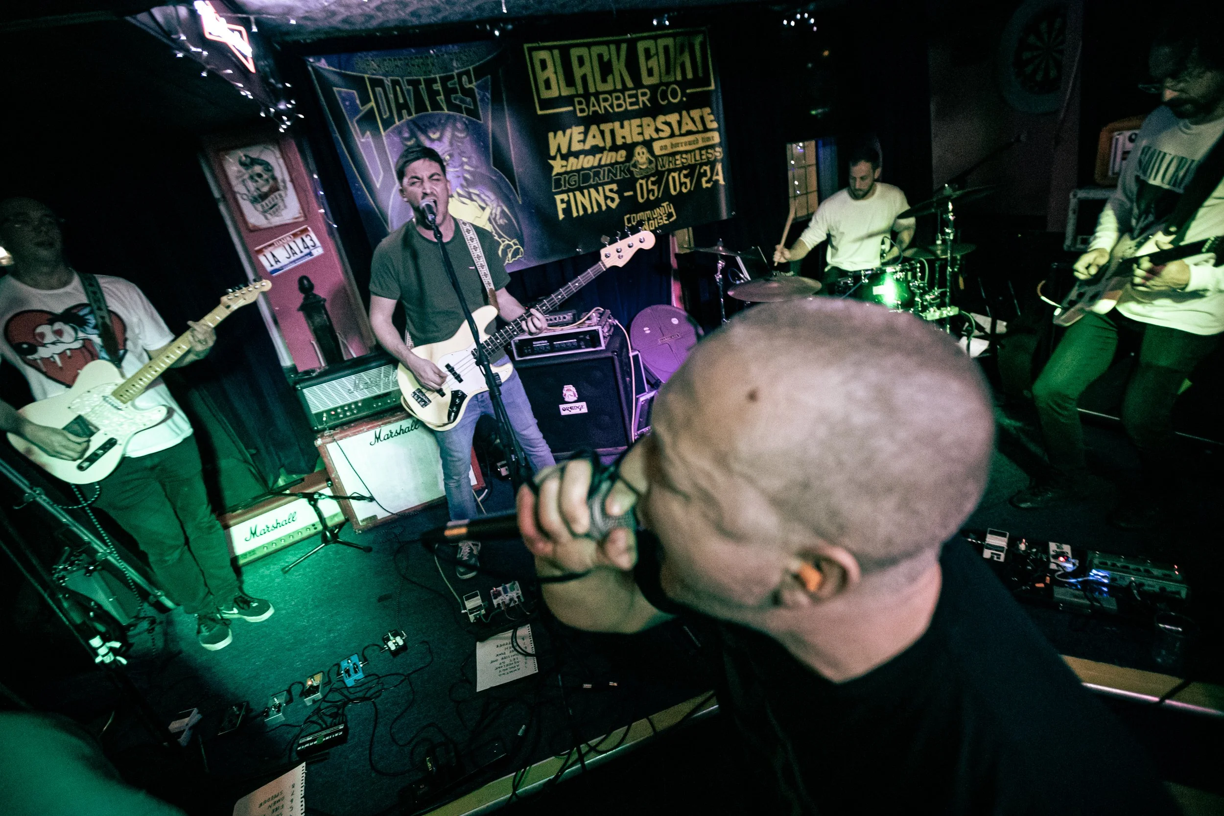 A live indoor band performance with four musicians on stage, including a vocalist with a microphone in the foreground, two guitarists, and a drummer. There are colorful banners and posters in the background.