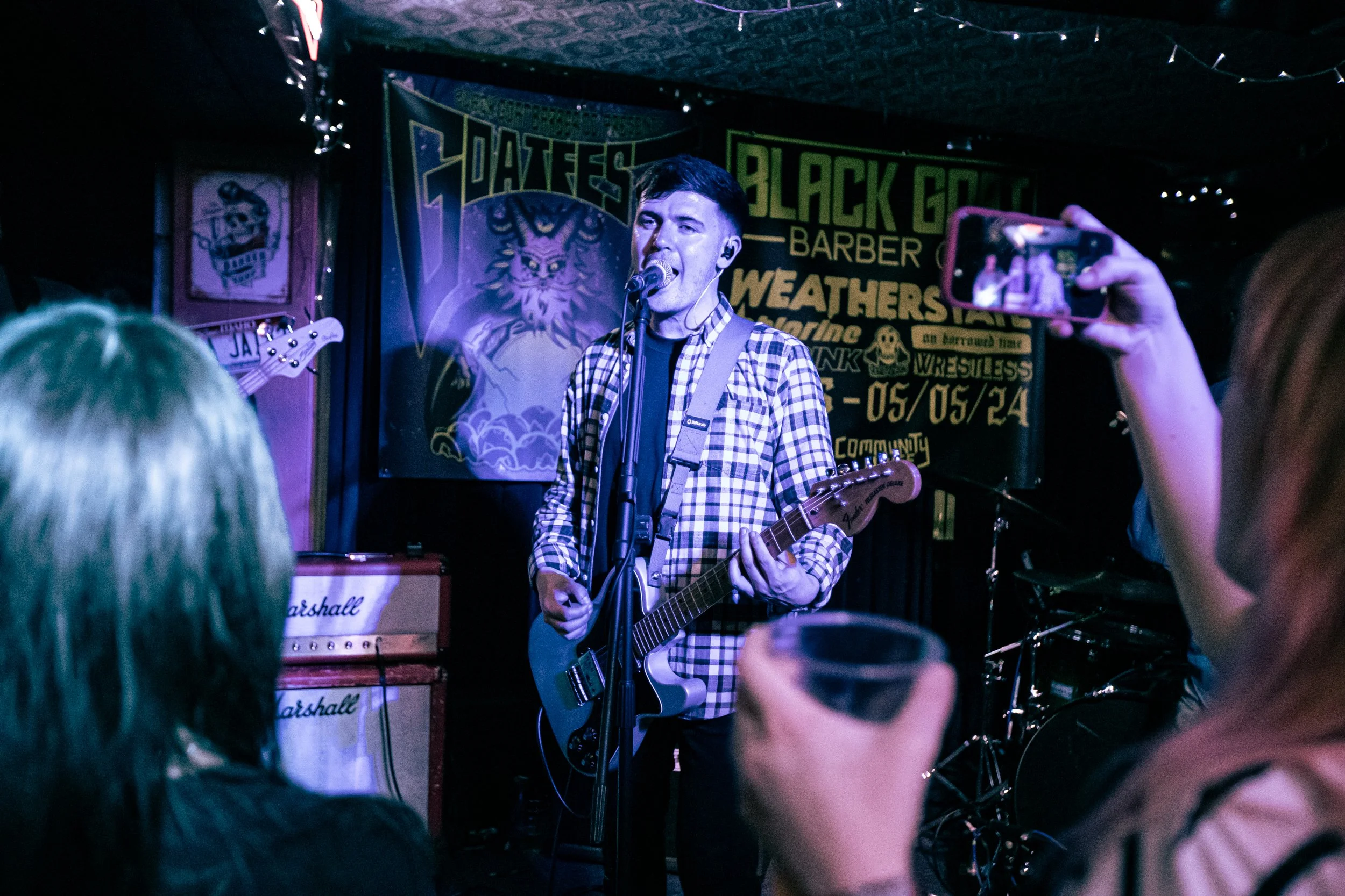 A male musician wearing a plaid shirt is singing and playing an electric guitar on stage in a dimly lit venue, with a person in the foreground taking a photo, and a black banner with yellow and green text hanging behind him.