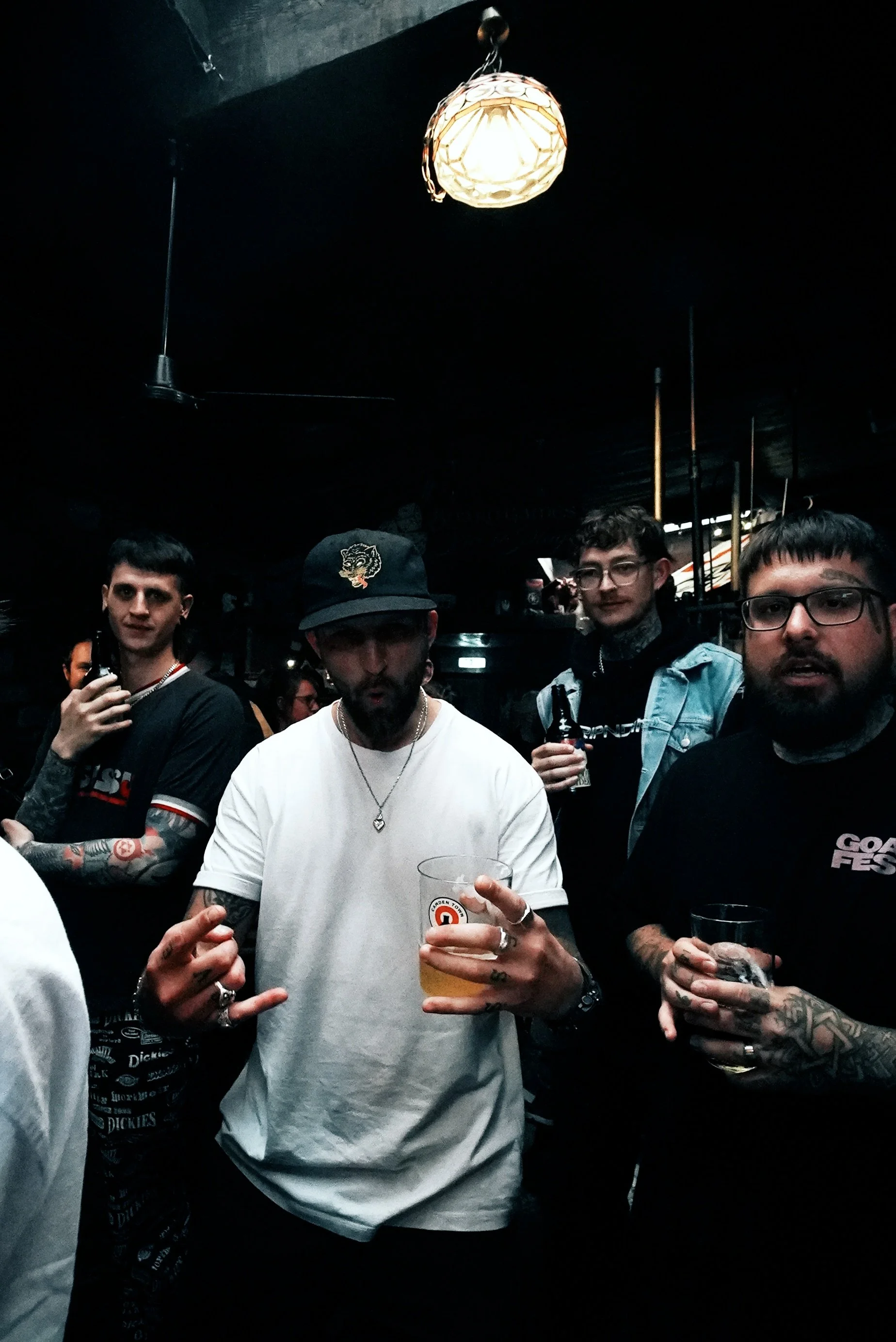 Group of four men in a dark indoor setting, some holding drinks, with one man in the center wearing a white t-shirt, black baseball cap, and jewelry. There is a hanging light fixture above them.