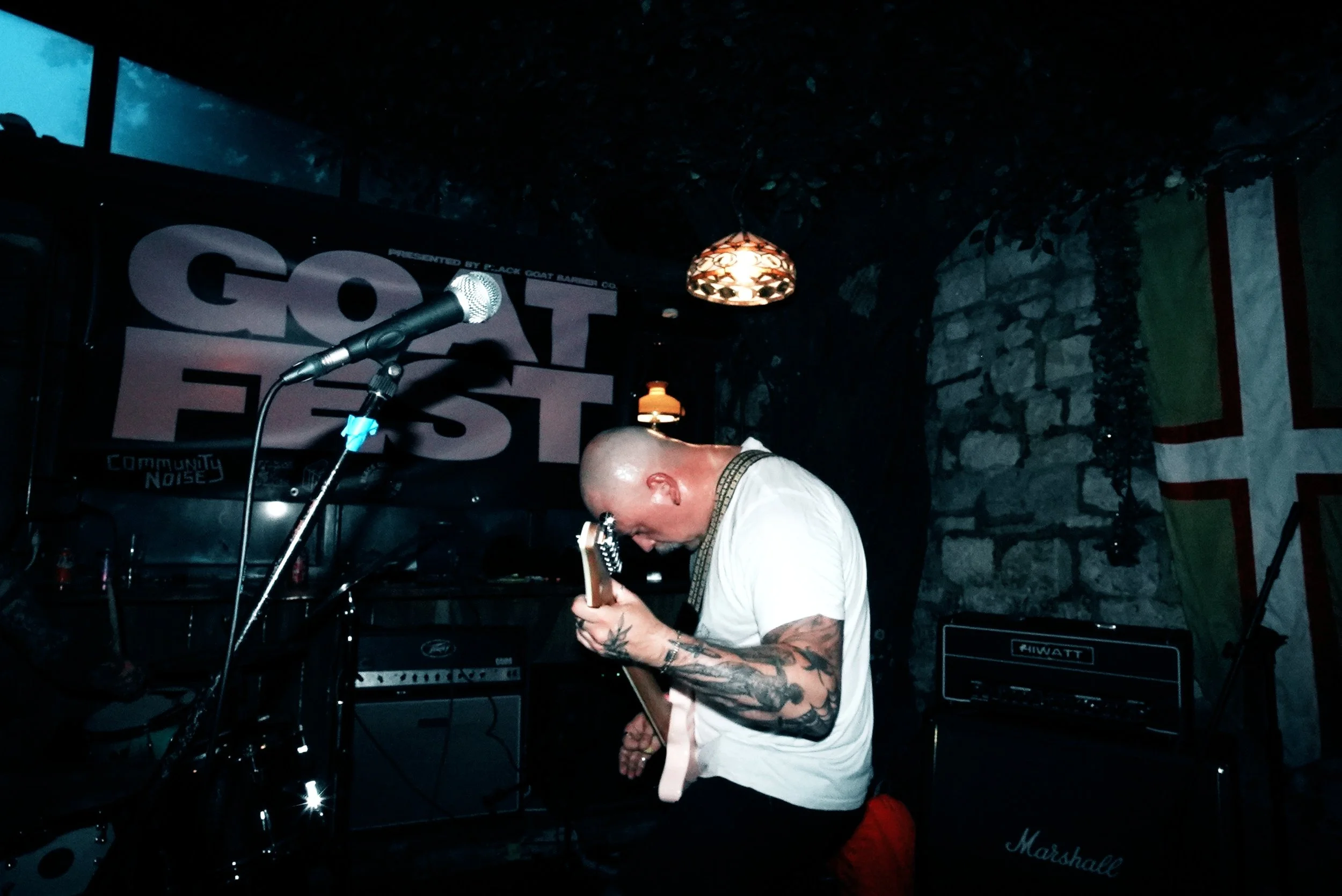 A musician with a bald head and tattoos on his arm is playing an electric guitar on a stage with a brick wall background. There is a microphone in front of him, and various amplifiers and equipment are visible. A banner with the words 'GOT FEST' hang