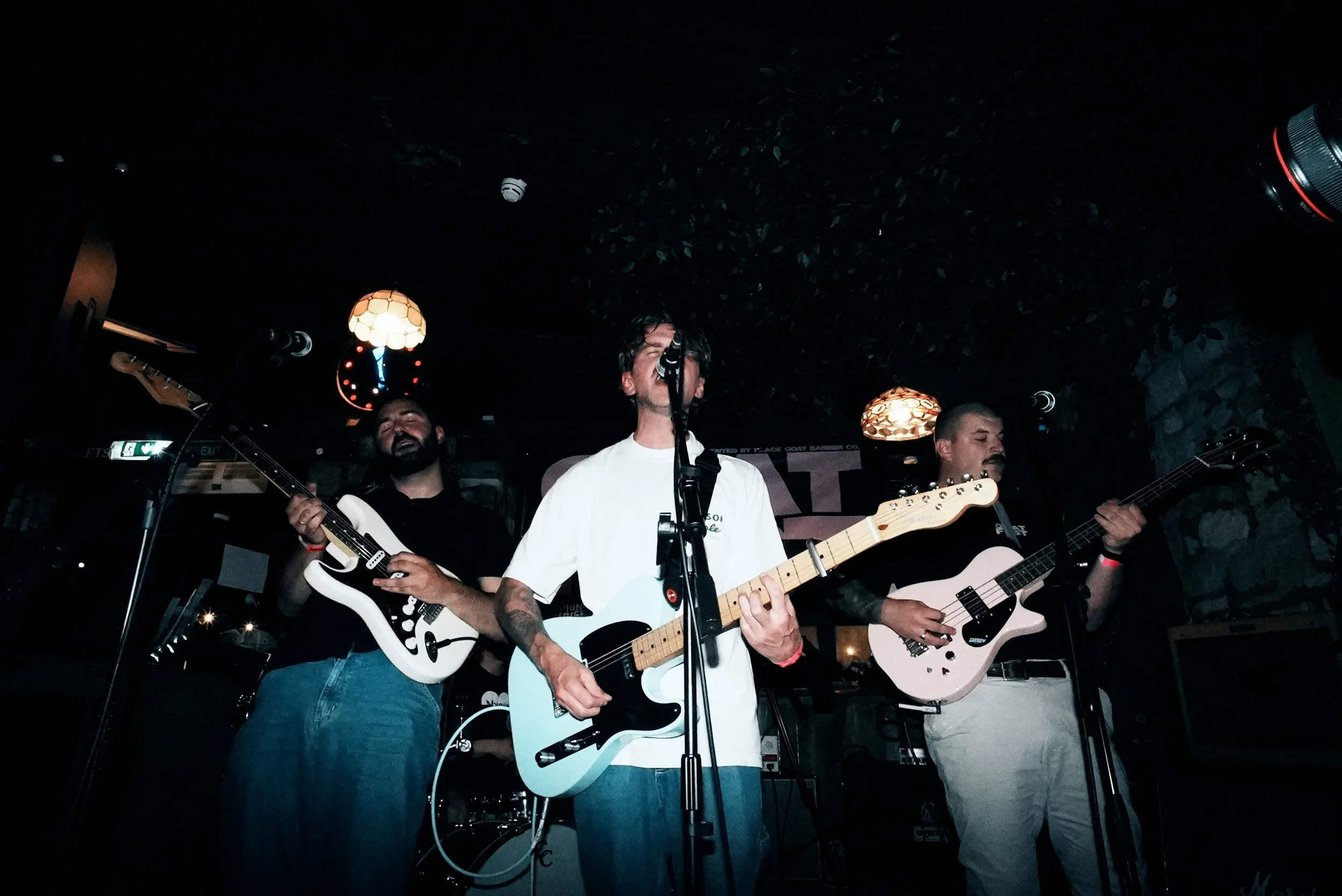 A band of three men performing live on stage with guitars and microphone, dimly lit with hanging lamps in the background.