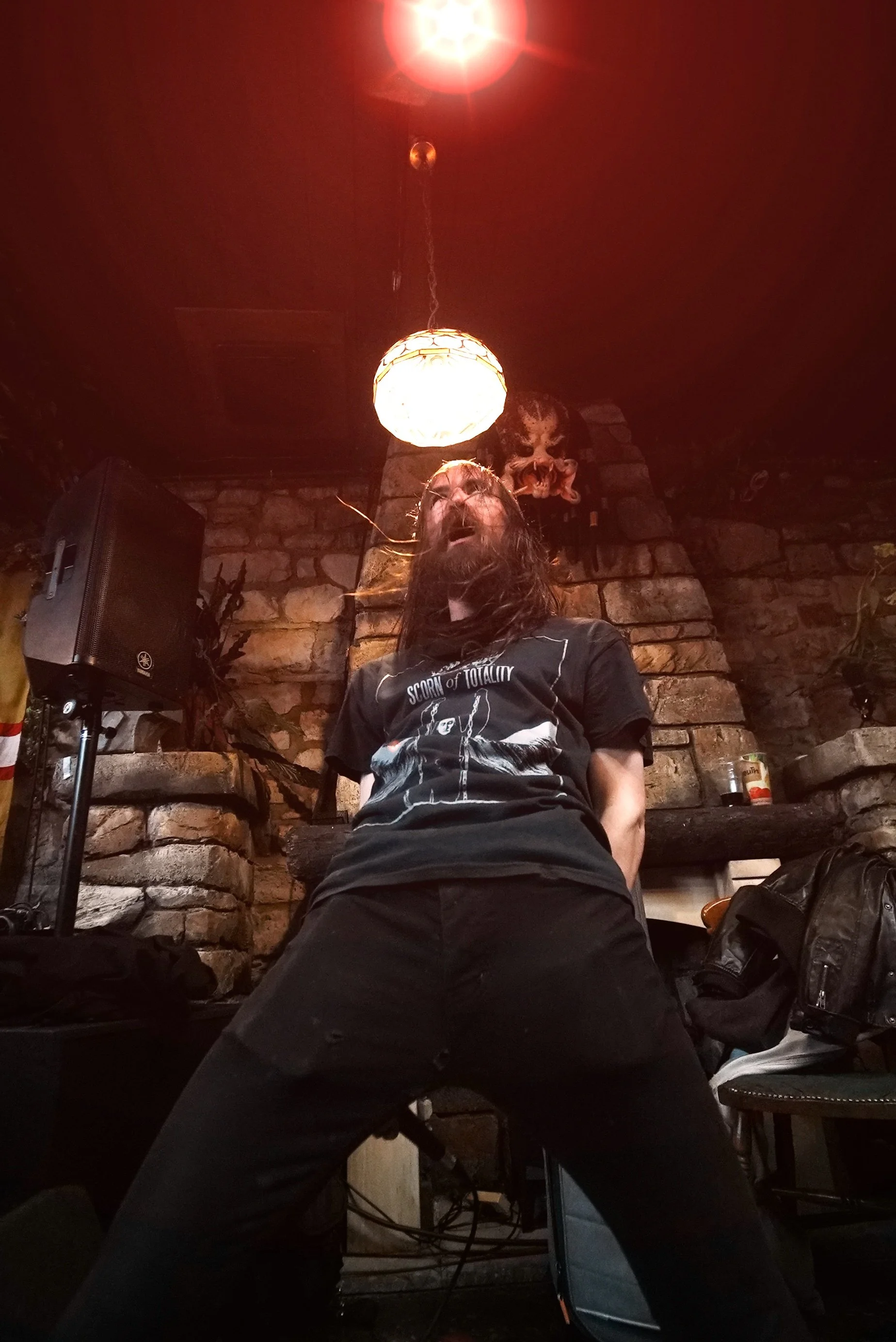 A person with long hair and a beard standing in front of a brick wall, wearing a black t-shirt with a graphic and text, in a dimly lit room with a red ceiling light and skull decoration.