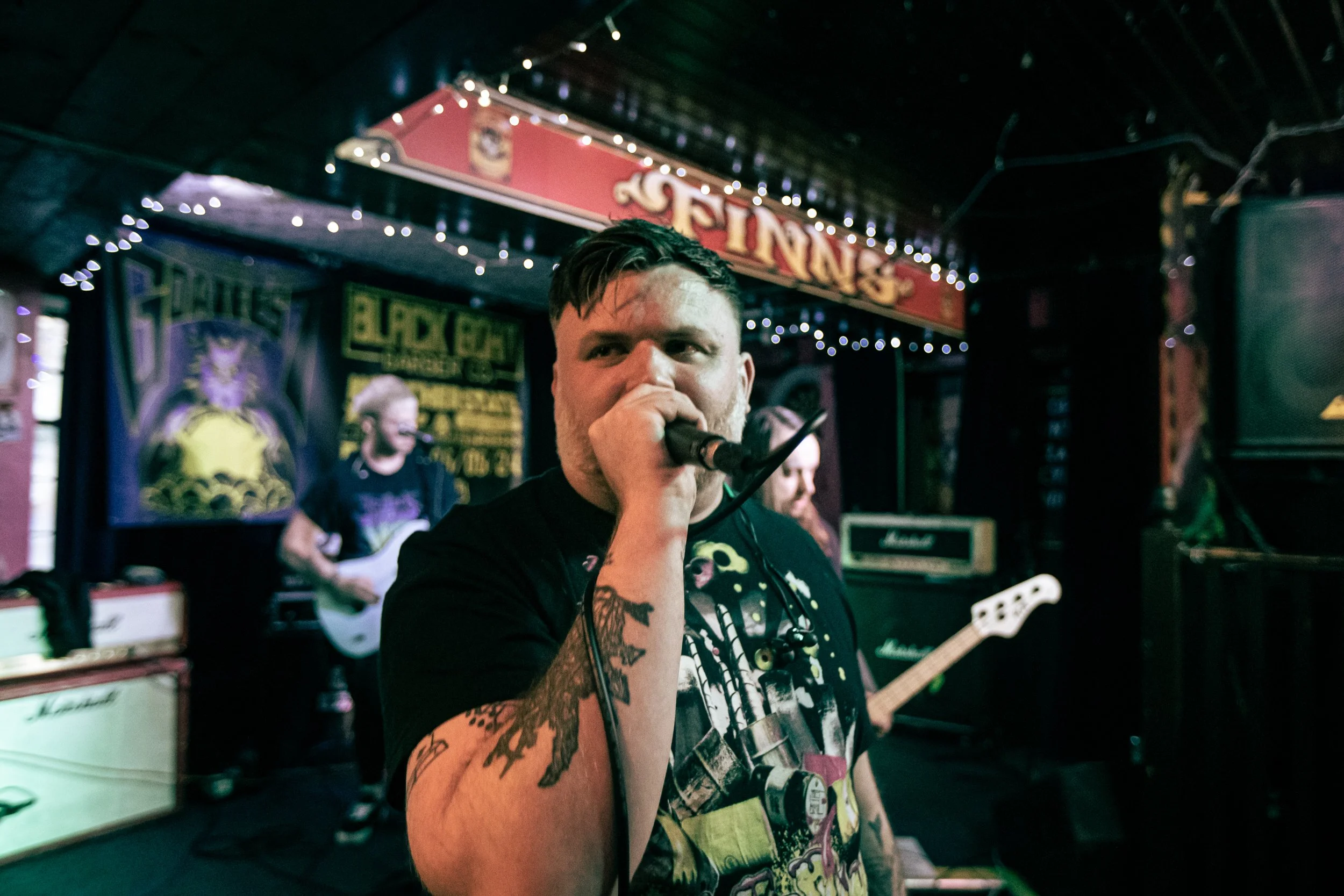 Male lead singer with tattoos on his arm holding a microphone on stage during a live music performance at a bar or venue called Fin's.