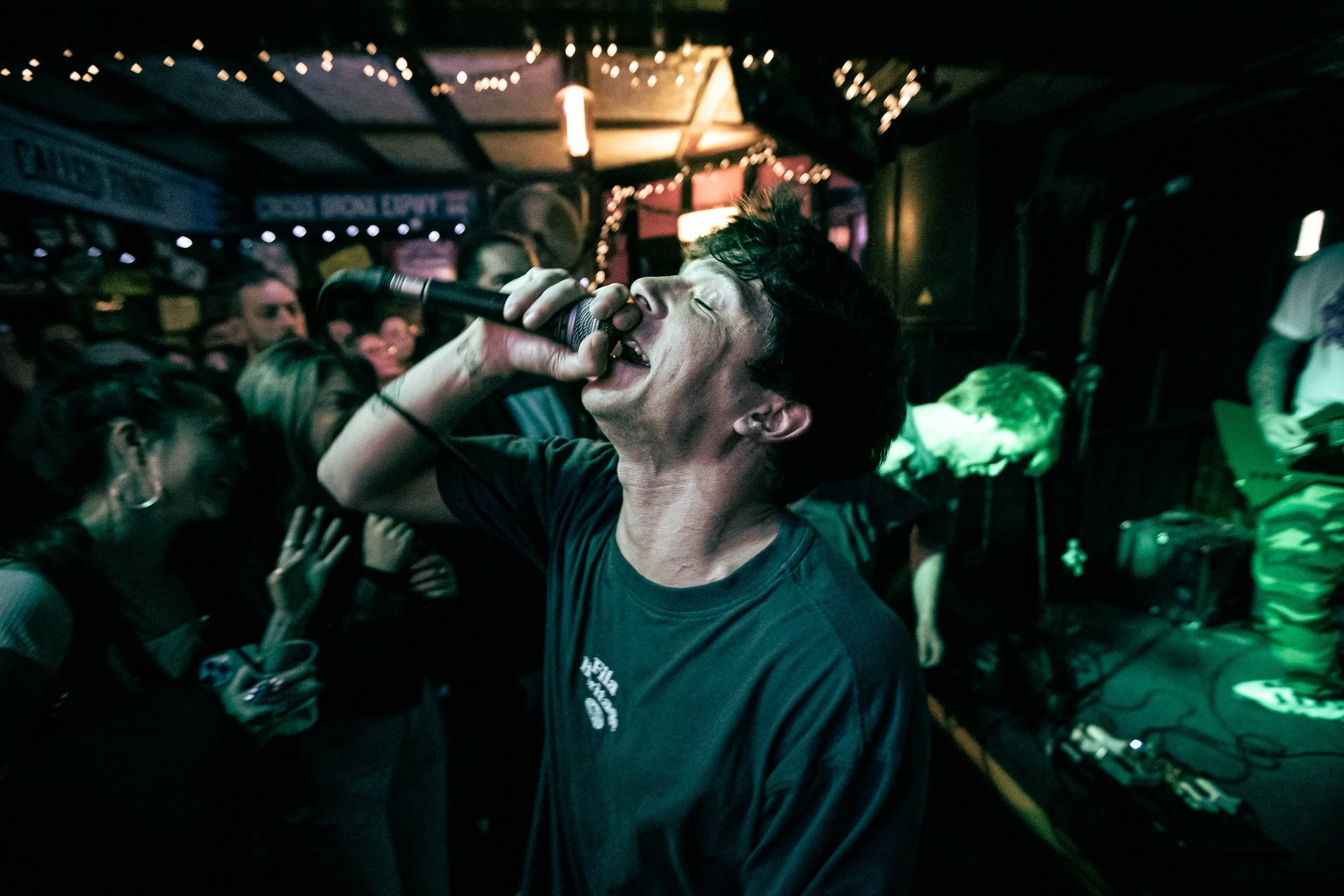 A man with dark hair singing into a microphone at a live music show, surrounded by a crowd in a dimly lit venue with string lights overhead.