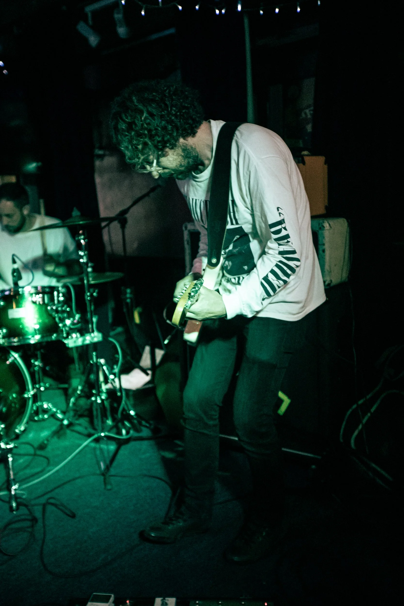 A man with curly hair and glasses playing an electric guitar on stage, with a drummer in the background, in a dimly lit music venue.