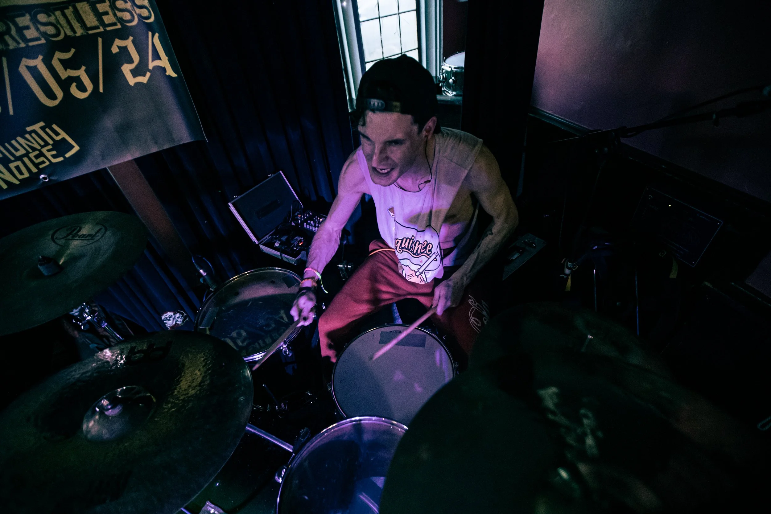 A young male drummer wearing a tank top, red pants, and a backwards baseball cap playing drums on stage with purple lighting.