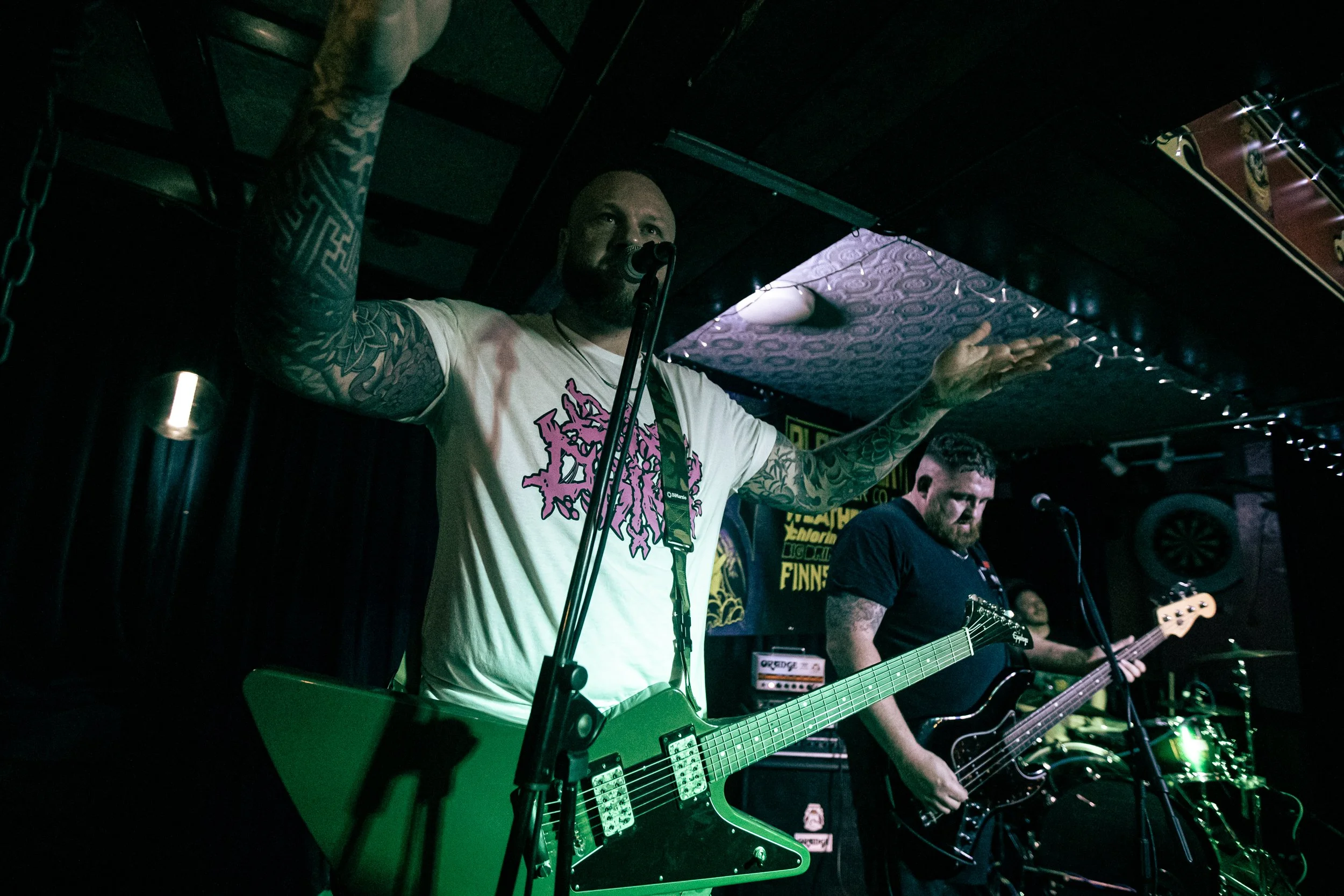 A band performs on stage with three members visible, one with tattoos and a white t-shirt singing into a microphone, playing a green electric guitar, with a black bass guitar and drummer in the background.