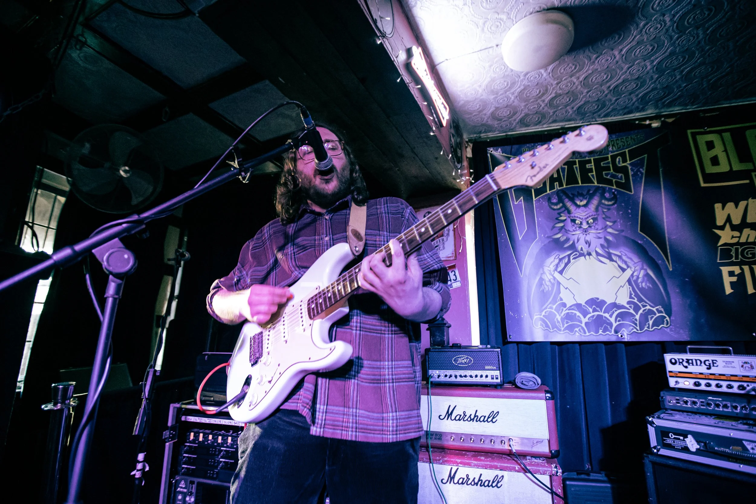A man singing and playing an electric guitar on stage at a music venue, with musical equipment around him and a colorful poster in the background.