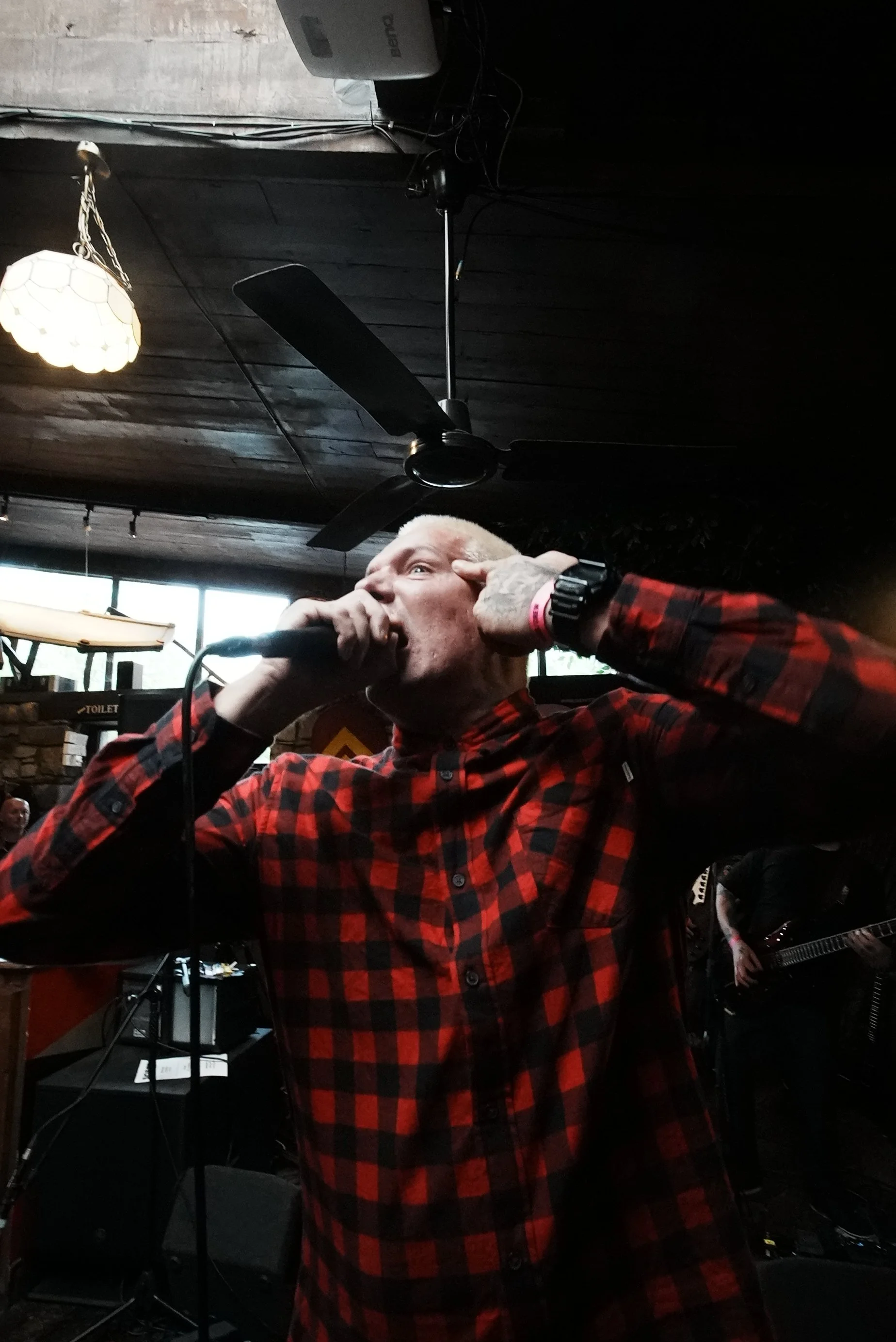A man with short blond hair singing into a microphone at a live music performance, wearing a red and black checkered shirt, with a band playing in the background.
