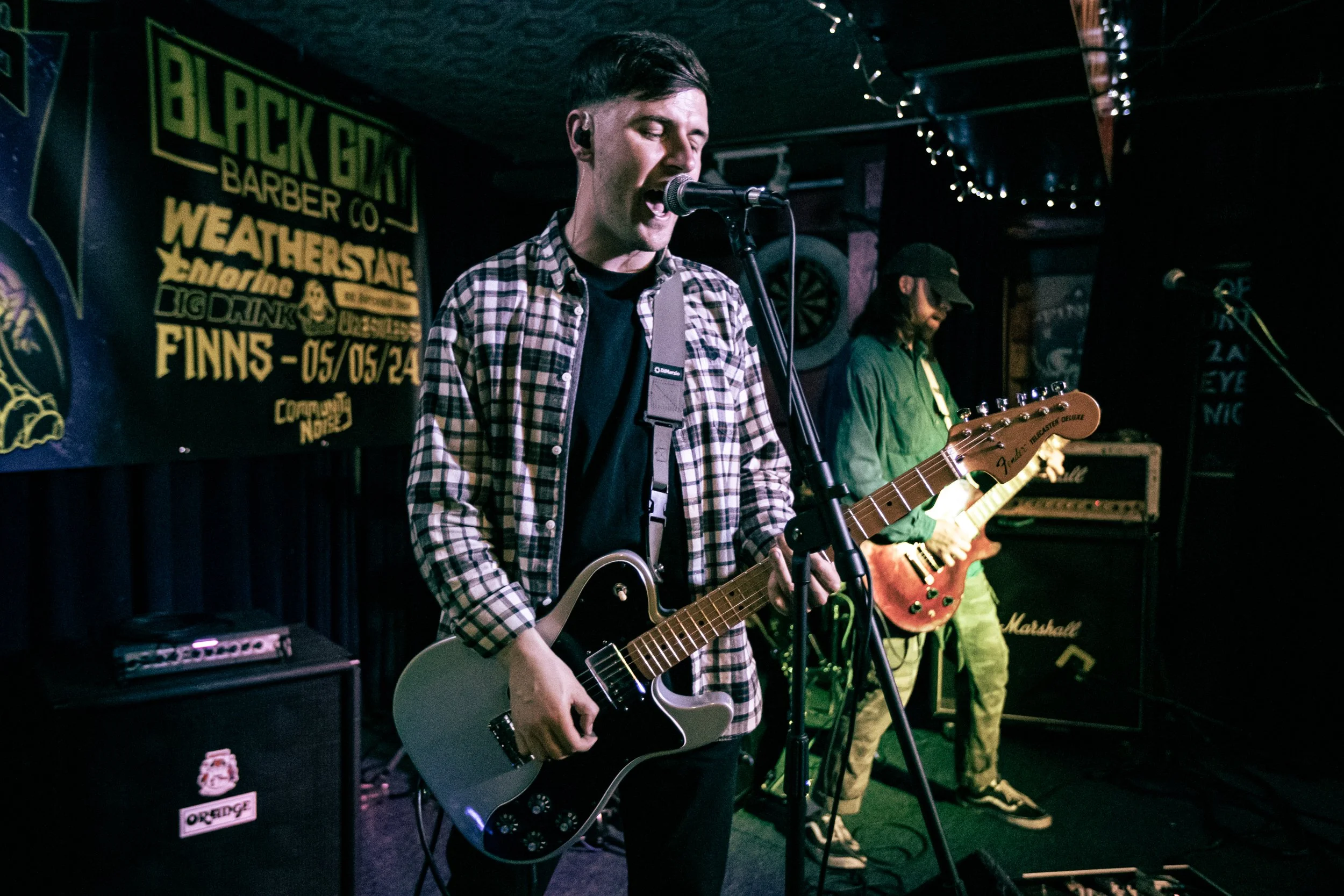 Two young male musicians performing on stage with guitars, one singing into a microphone, in a dimly lit venue with promotional posters on the wall.