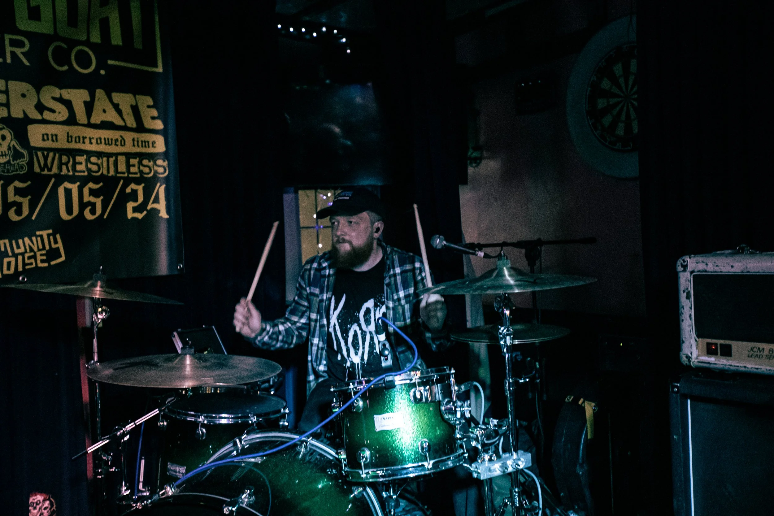 A man with a beard and cap playing a green drum set on stage with a microphone nearby, in a dark venue with a banner and dartboard on the wall.