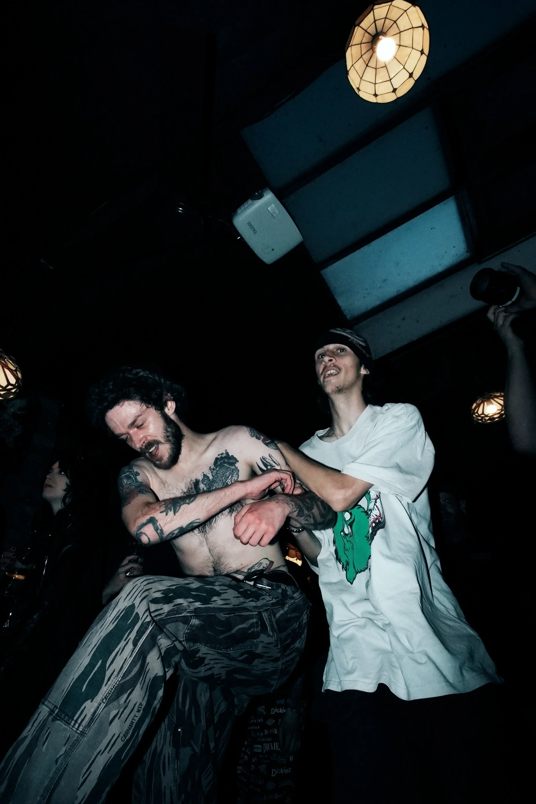 Two shirtless men, one with tattoos and dark hair, and another wearing a white T-shirt with a green graphic, are in a dark room with hanging lamps. The man with dark hair appears to be in discomfort or pain, while the other stands behind him. Someone