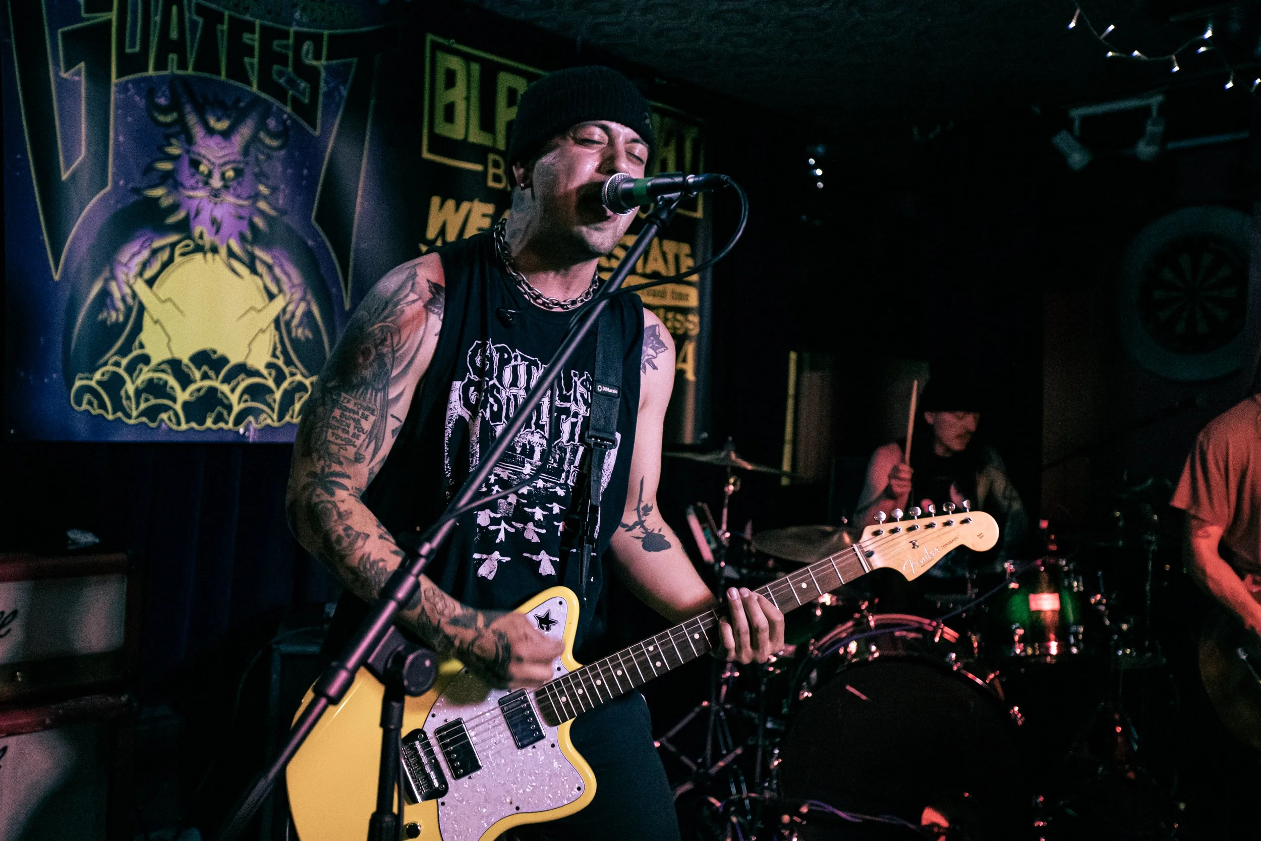 A musician with tattoos, wearing a black beanie and sleeveless shirt, plays a yellow electric guitar and sings into a microphone during a live performance in a dimly lit venue with a band behind him and signs in the background.