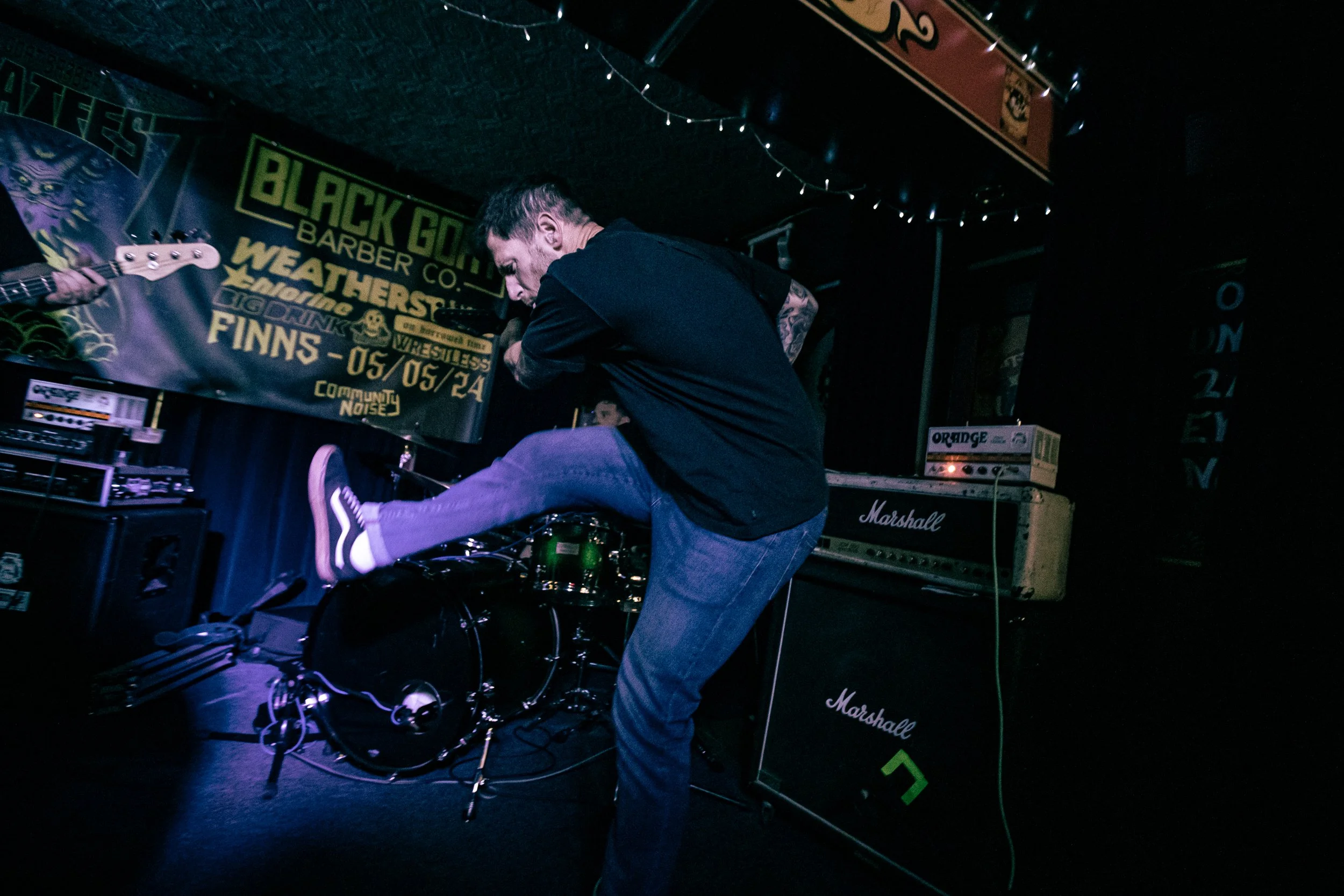 A musician performs on stage at a concert, with a black drum set in front and amplifiers behind. The stage has a banner with event details and various musical equipment around.