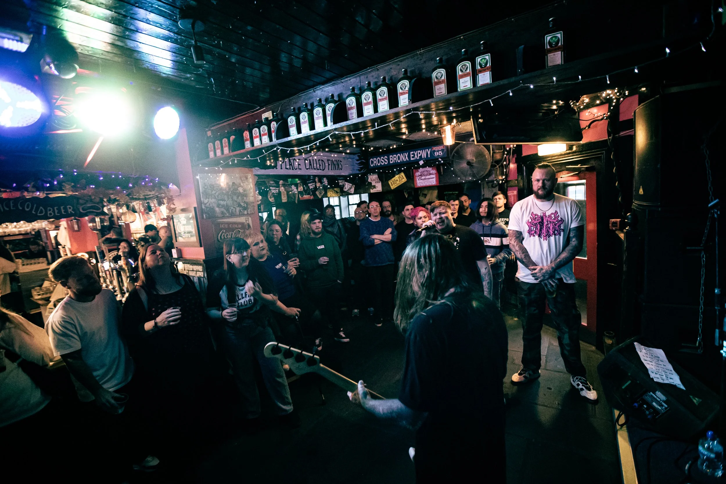Live music performance at a bar or venue with a guitarist playing in front of an audience, including a singer and several spectators, with dim lighting and colorful stage lights.
