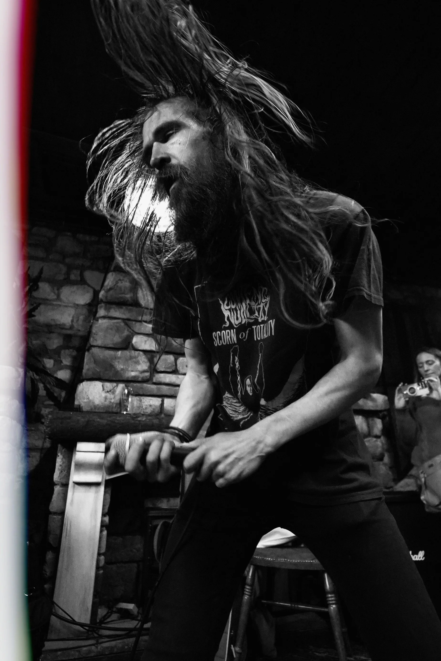 A man with long hair and a beard passionately singing or performing on stage, holding a microphone, in a dimly lit environment with a brick wall background.