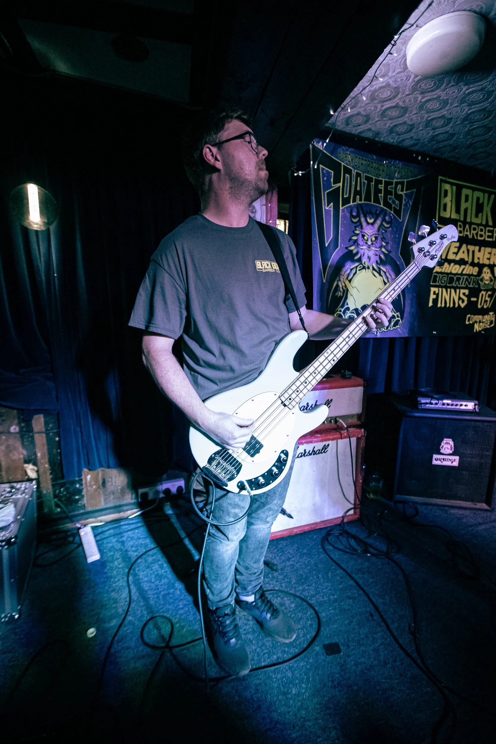 A man wearing glasses and a gray t-shirt playing a white bass guitar on stage at a music venue.