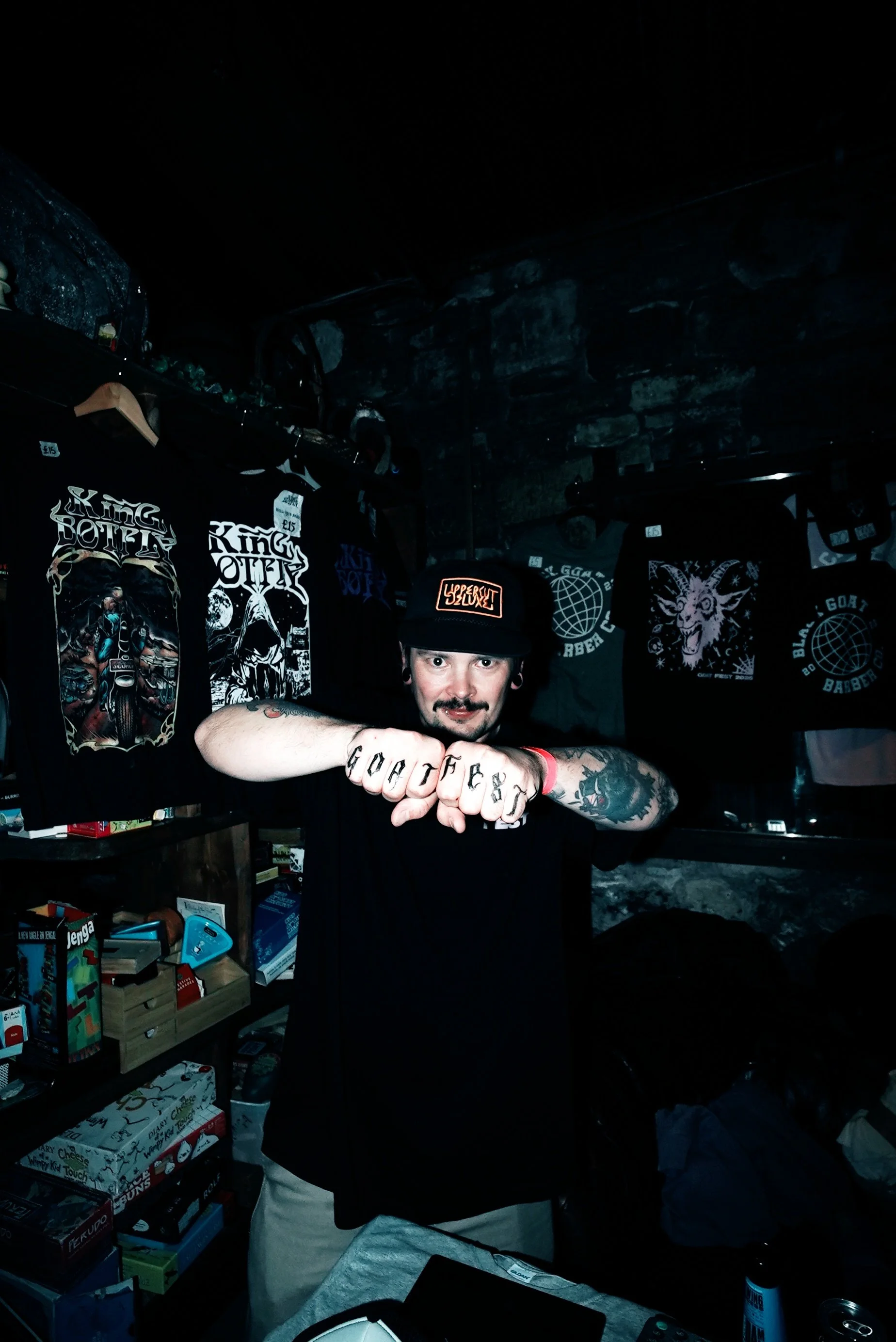 A man wearing a black cap and black T-shirt, standing indoors, showing his fists with tattoos reading 'GOAT FEST'. The background includes black T-shirts with graphic prints hanging on the wall, and shelves with board games and boxed items.