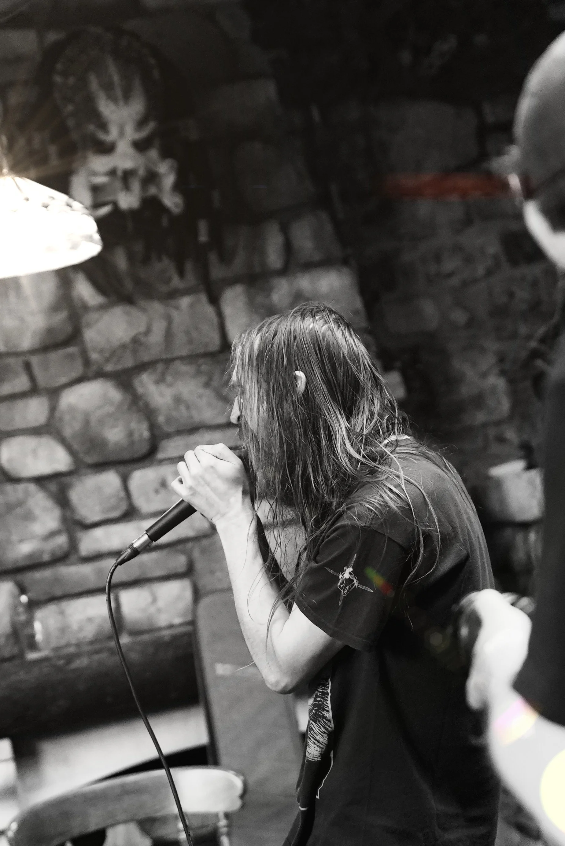 A person with long hair singing into a microphone in a dimly lit room with a stone wall background.