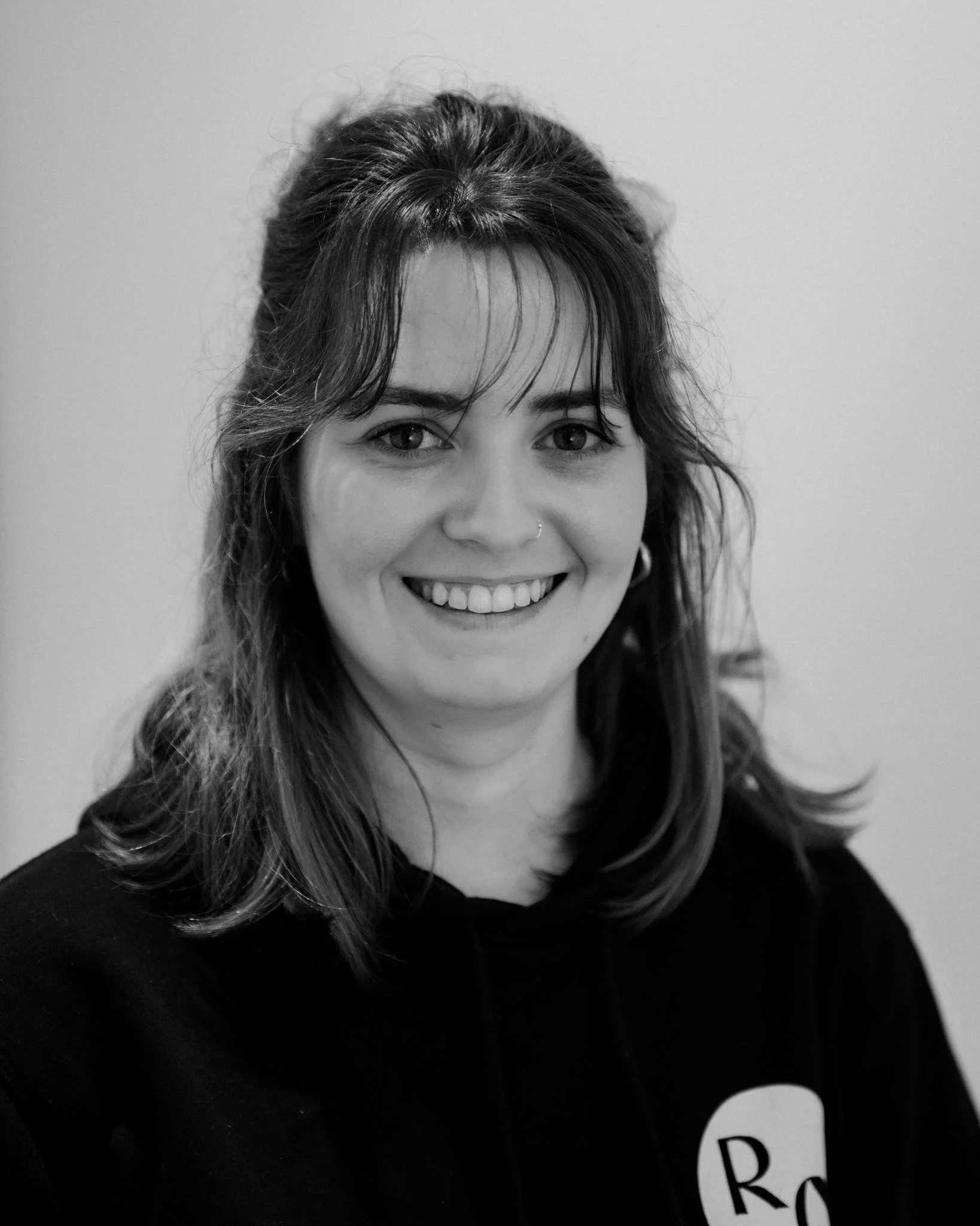 Black and white portrait of a smiling woman with shoulder-length hair, nose piercing, and wearing a hoodie with the initials 'RC' on it.