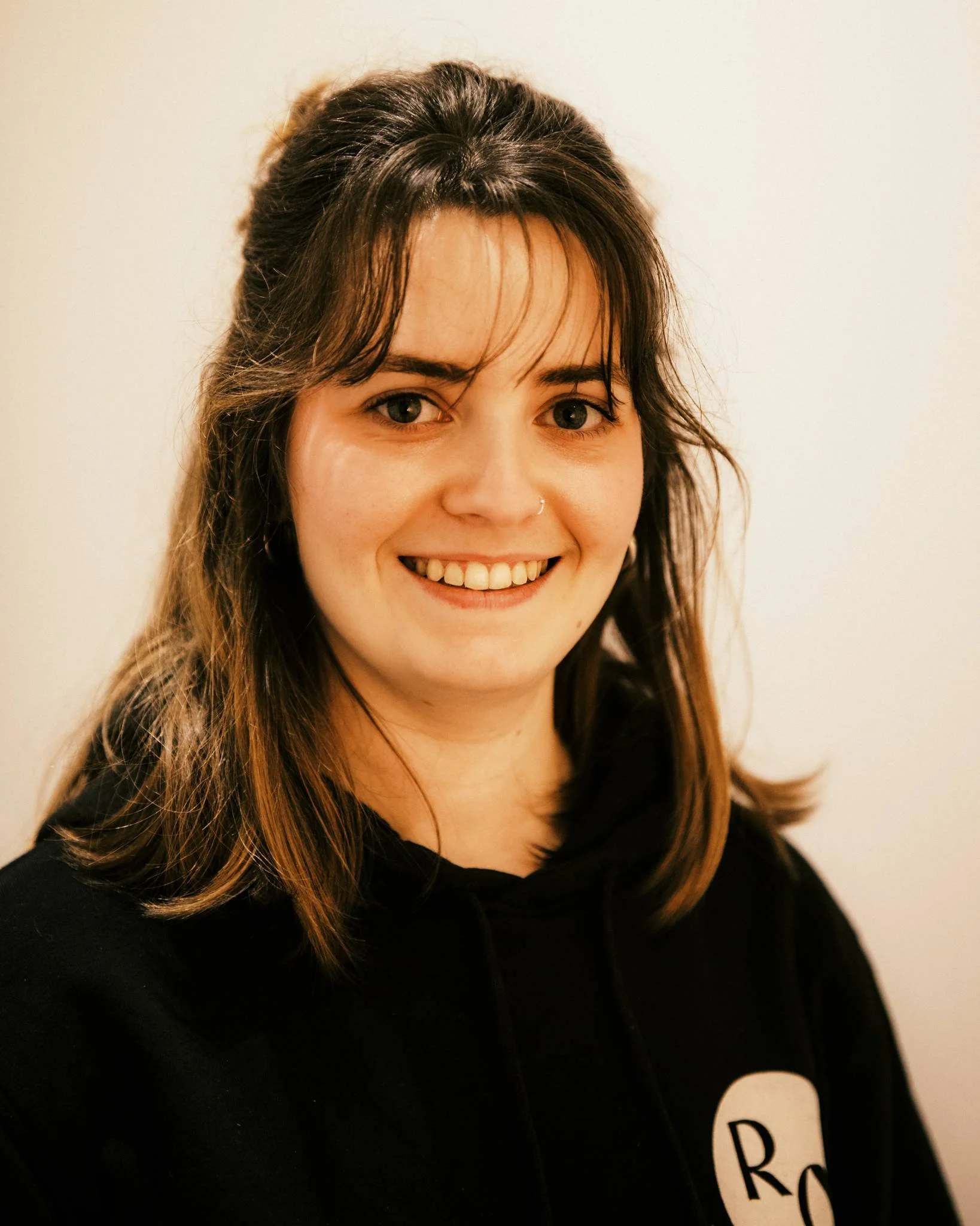 A woman with shoulder-length brown hair, a nose ring, and a black hoodie smiling against a plain beige wall.