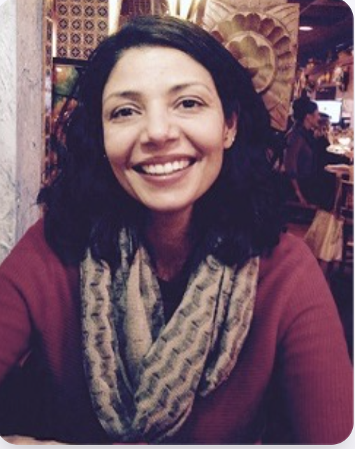 A woman with dark, wavy hair and a wide smile, wearing a maroon top and a patterned scarf, sitting in a warmly lit restaurant or cafe.