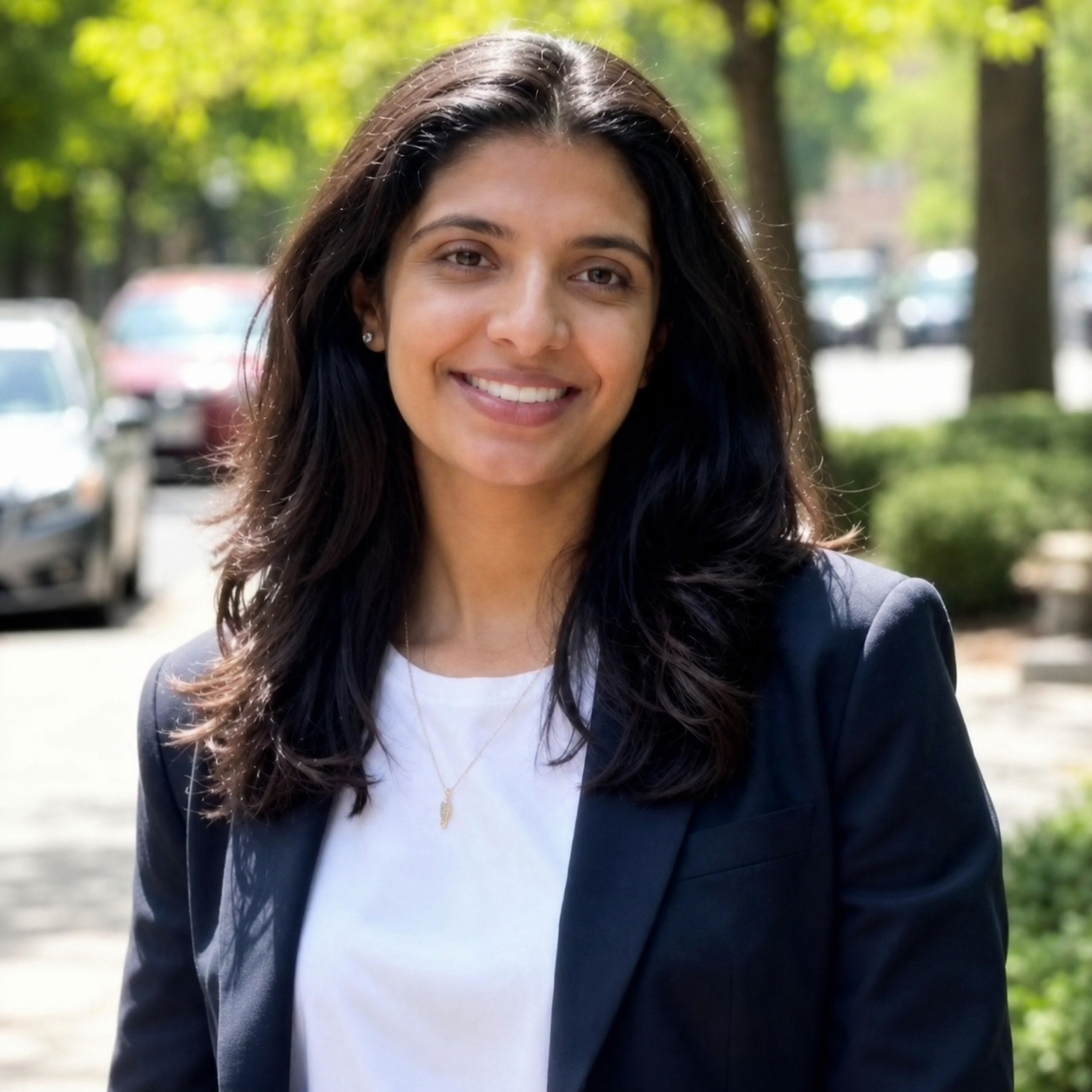 A woman with long dark hair, wearing a black blazer and white shirt, smiling outdoors with trees and parked cars in the background.