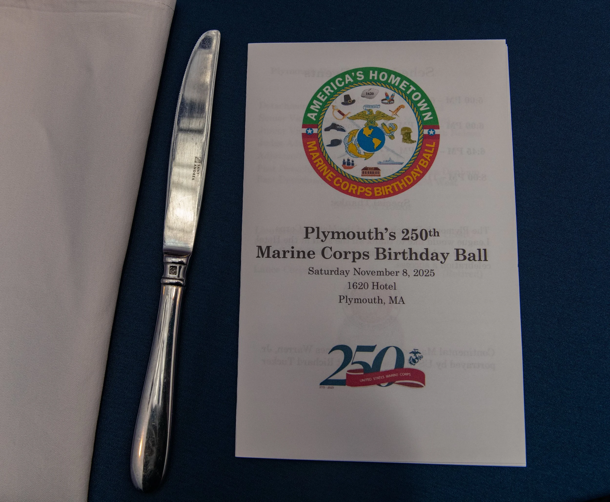 Event program for Plymouth's 250th Marine Corps Birthday Ball, dated Saturday, November 8, 2025, at the 1620 Hotel in Plymouth, MA, accompanied by a silver knife on a table with a blue tablecloth.