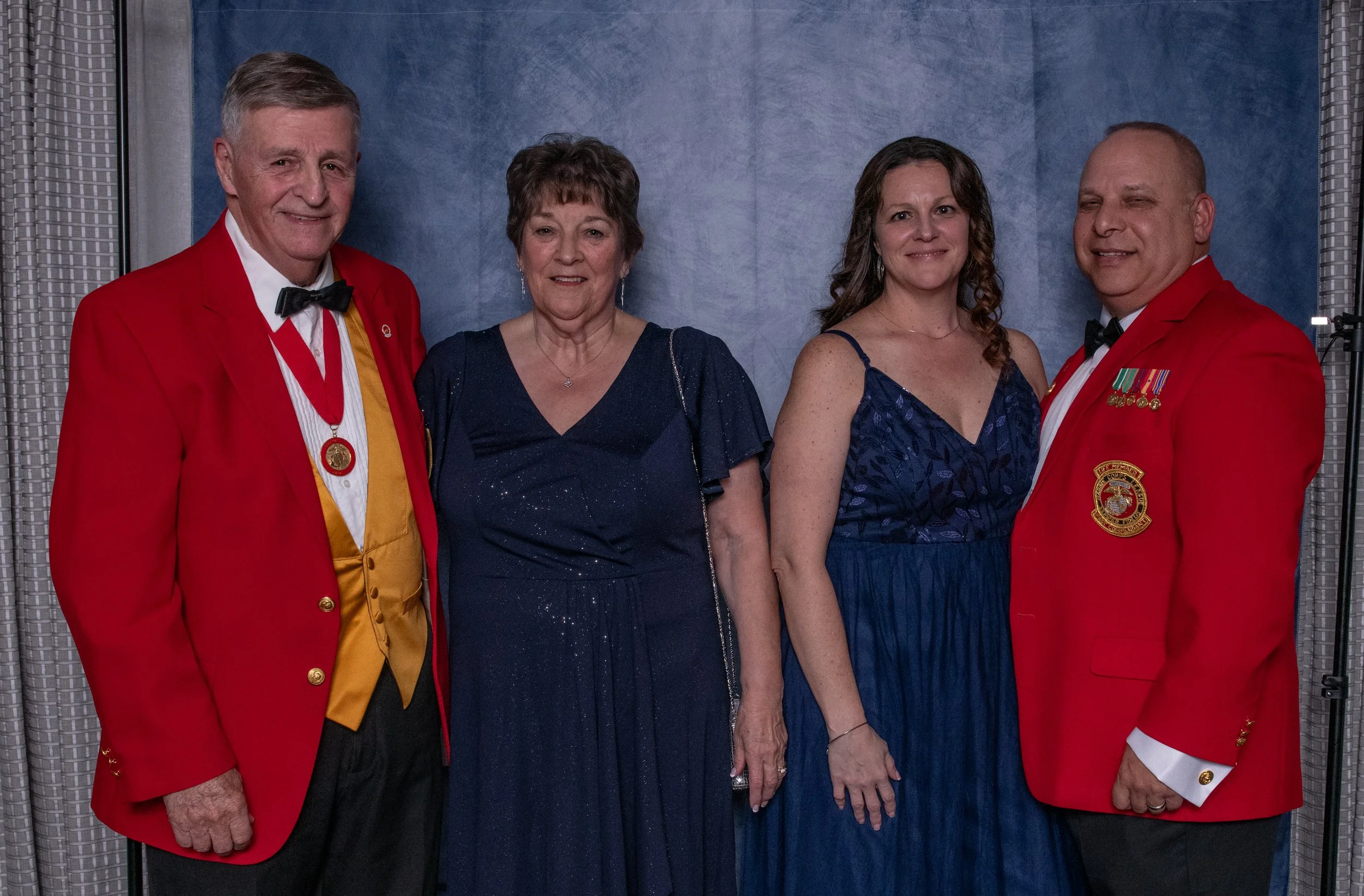 Four adults, two men and two women, standing together at a formal event with a blue backdrop. The men are dressed in red blazers with medals, while the women wear elegant blue dresses.