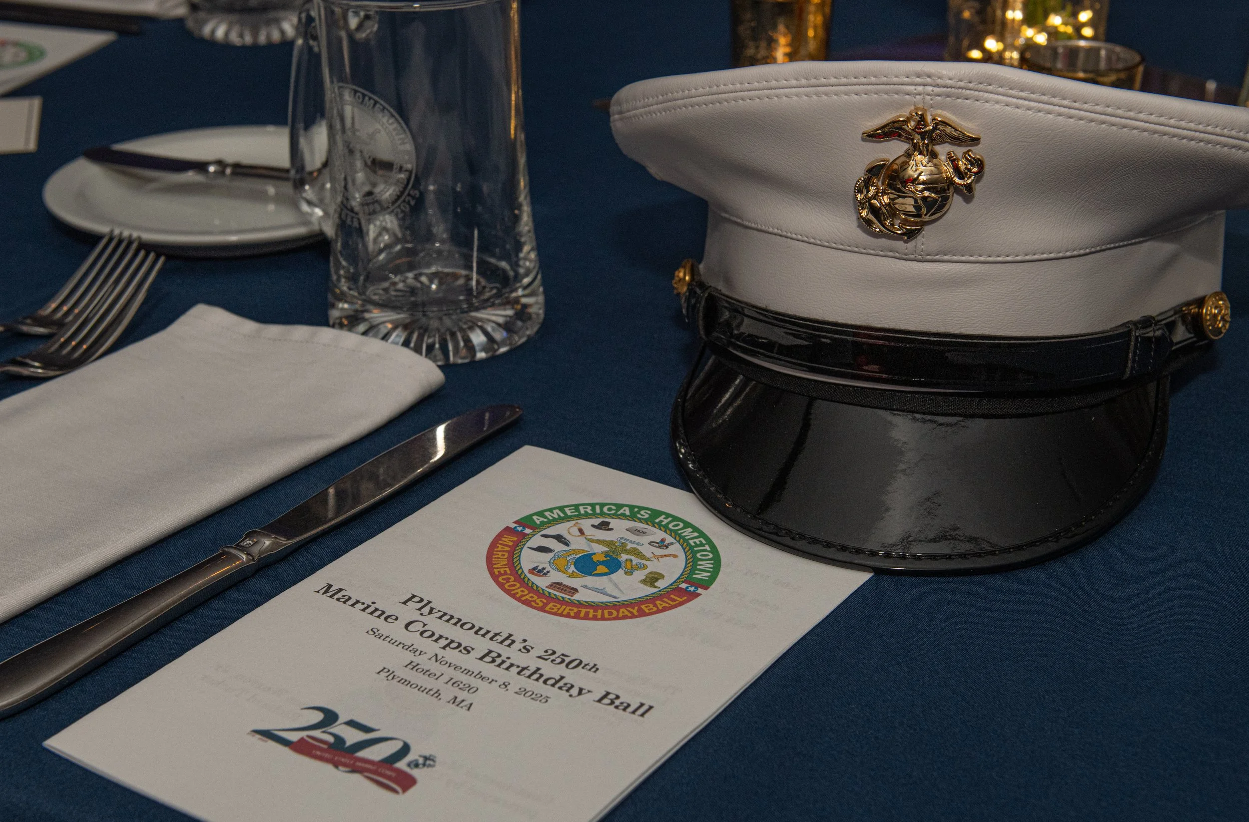 A table setup with a white cap with a Marine Corps emblem, a menu for Plymouth’s 250th Marine Corps Birthday Ball, a glass of water, a white napkin, silverware, and a clear glass. The menu features a Marine Corps emblem and colorful decoration, celeb