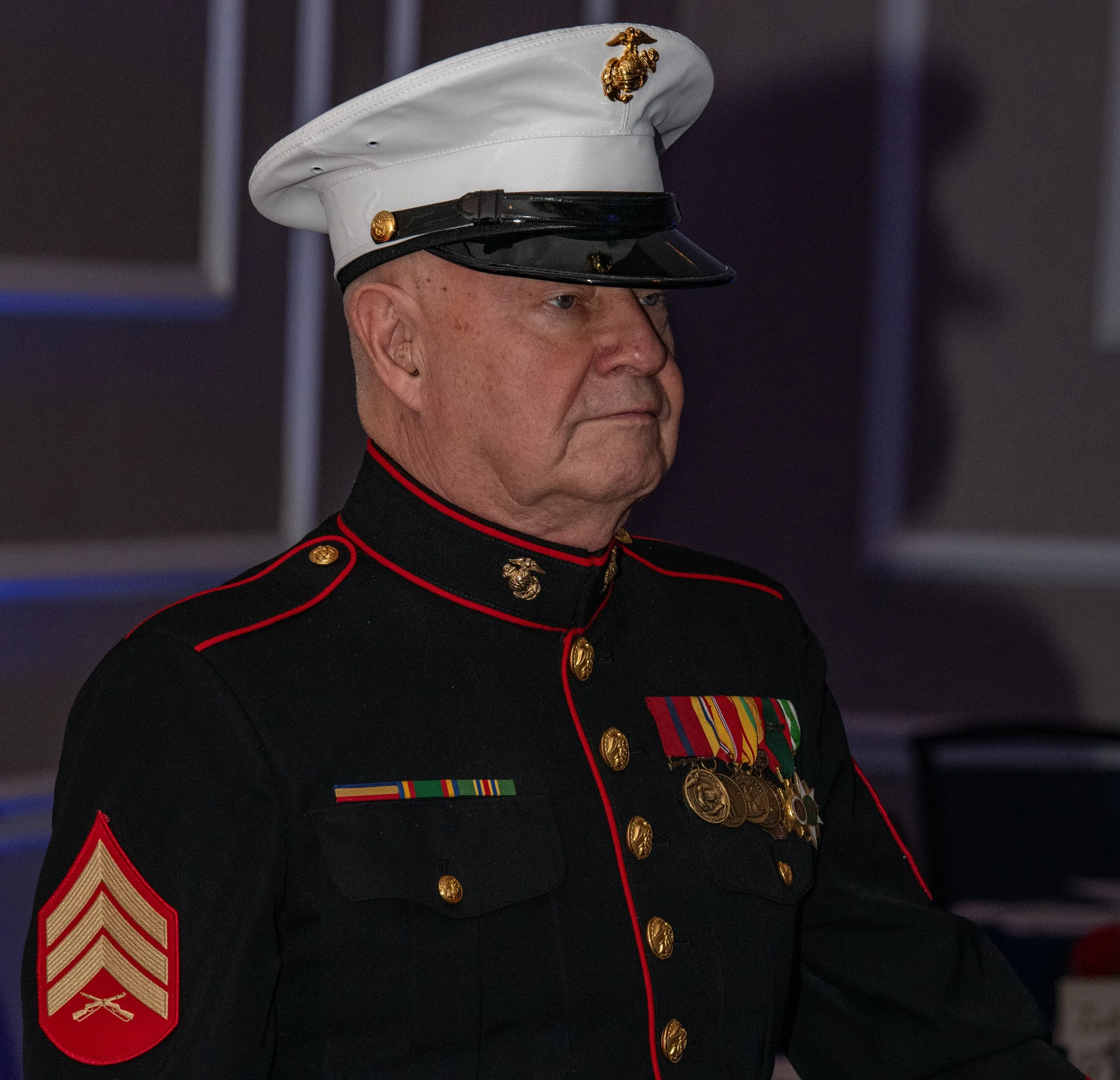A United States Marine in dress uniform, wearing a white cap with a gold emblem, a dark jacket with red piping, and medals on the chest.
