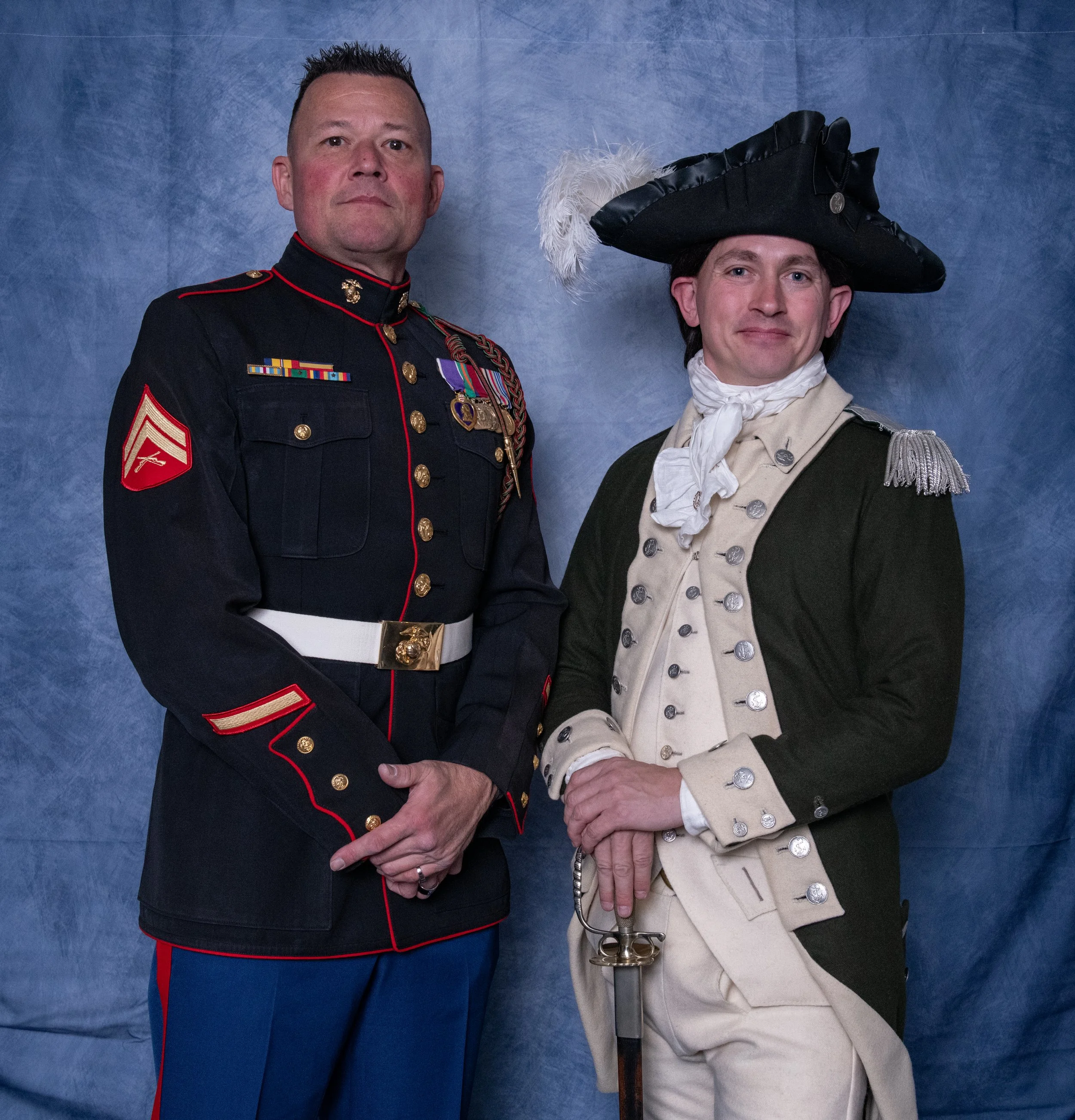 Two men dressed in historical military uniforms standing against a blue backdrop.