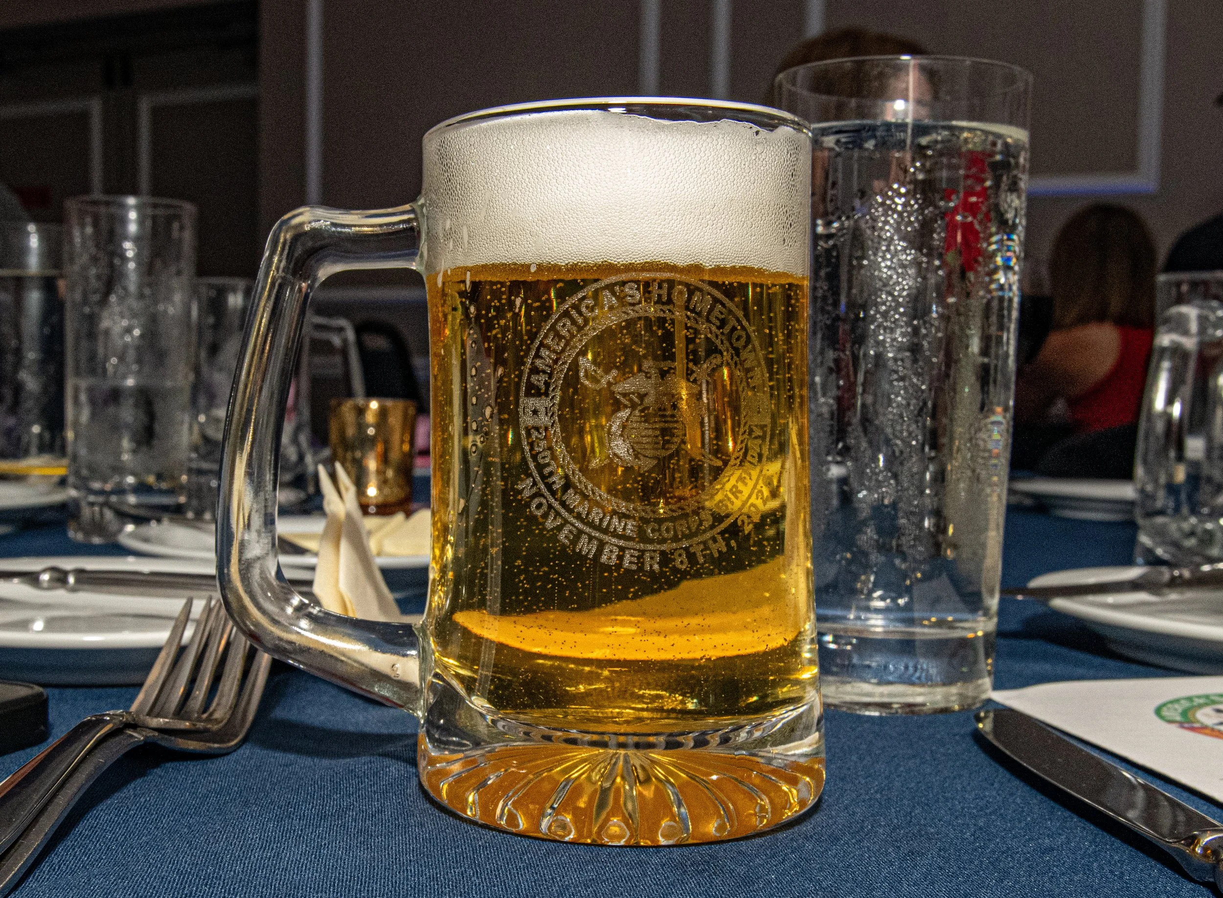 A glass mug of beer with a foamy head on a table, with other glasses and tableware around, in a restaurant setting.