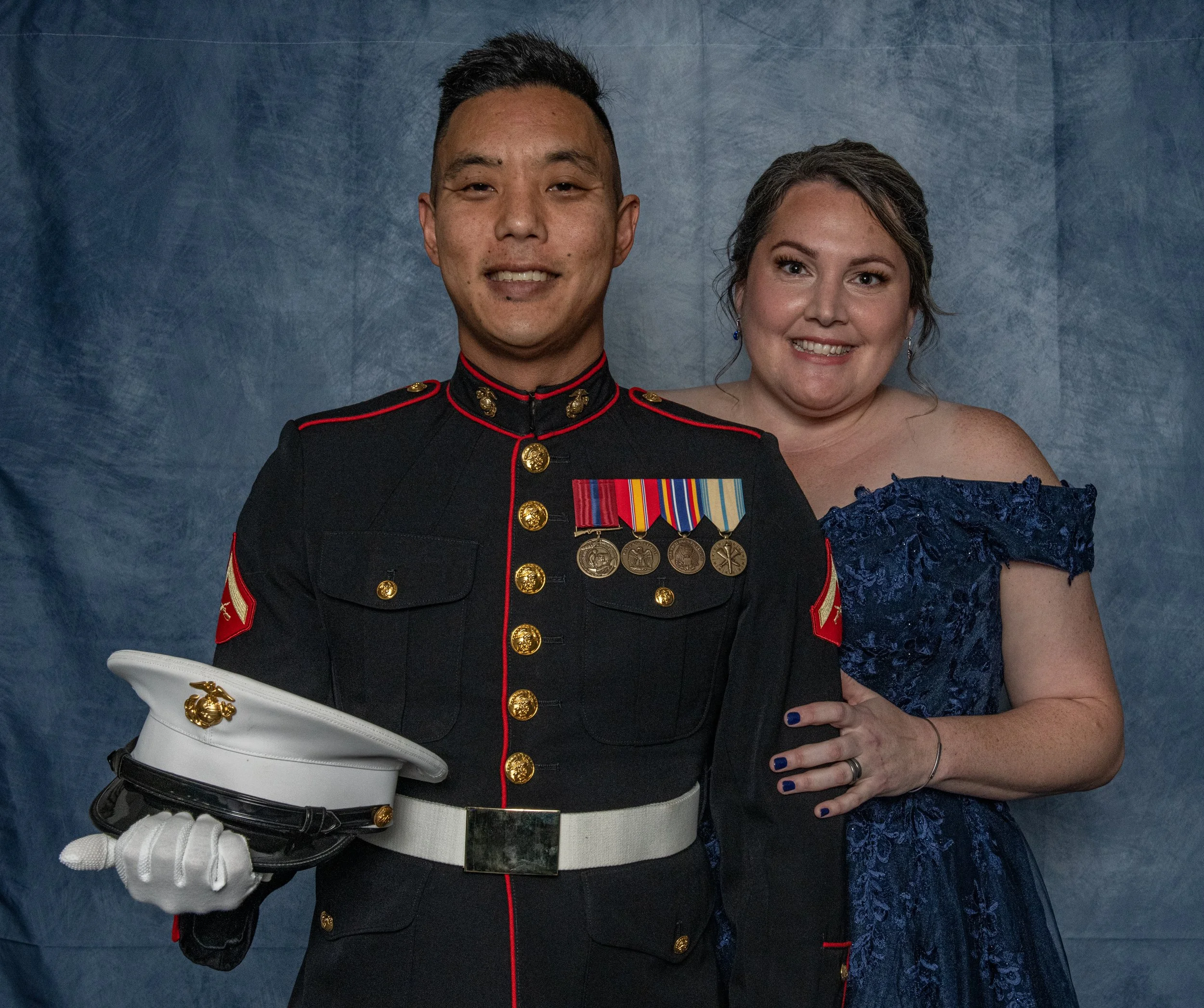 A person in a United States Marine Corps dress uniform standing next to a woman in a blue off-shoulder dress, both smiling for the camera against a blue background.