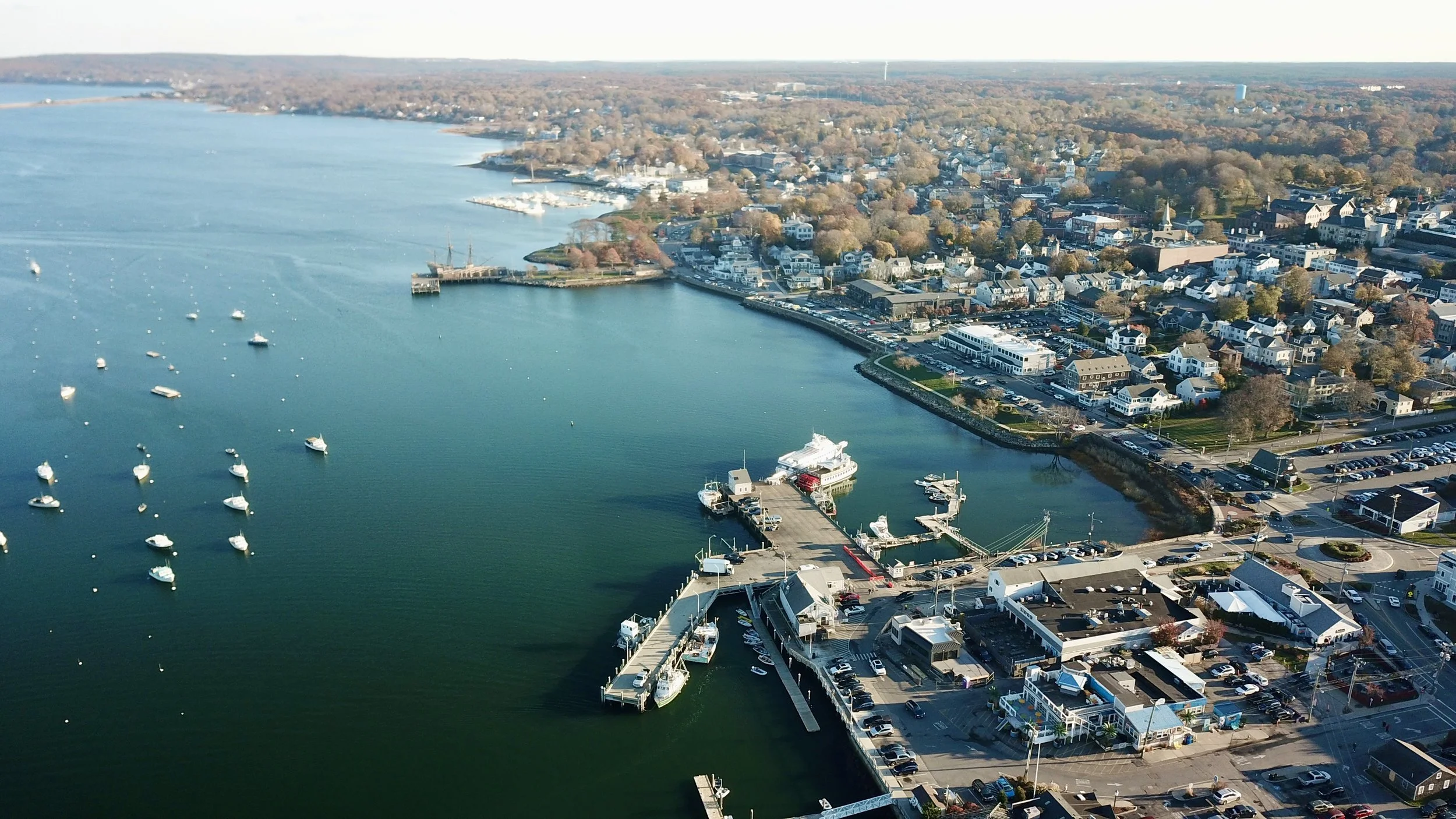 Aerial view of a marina with boats docked, adjacent to a coastal town with numerous buildings, streets, and parking lots, overlooking a large body of water.