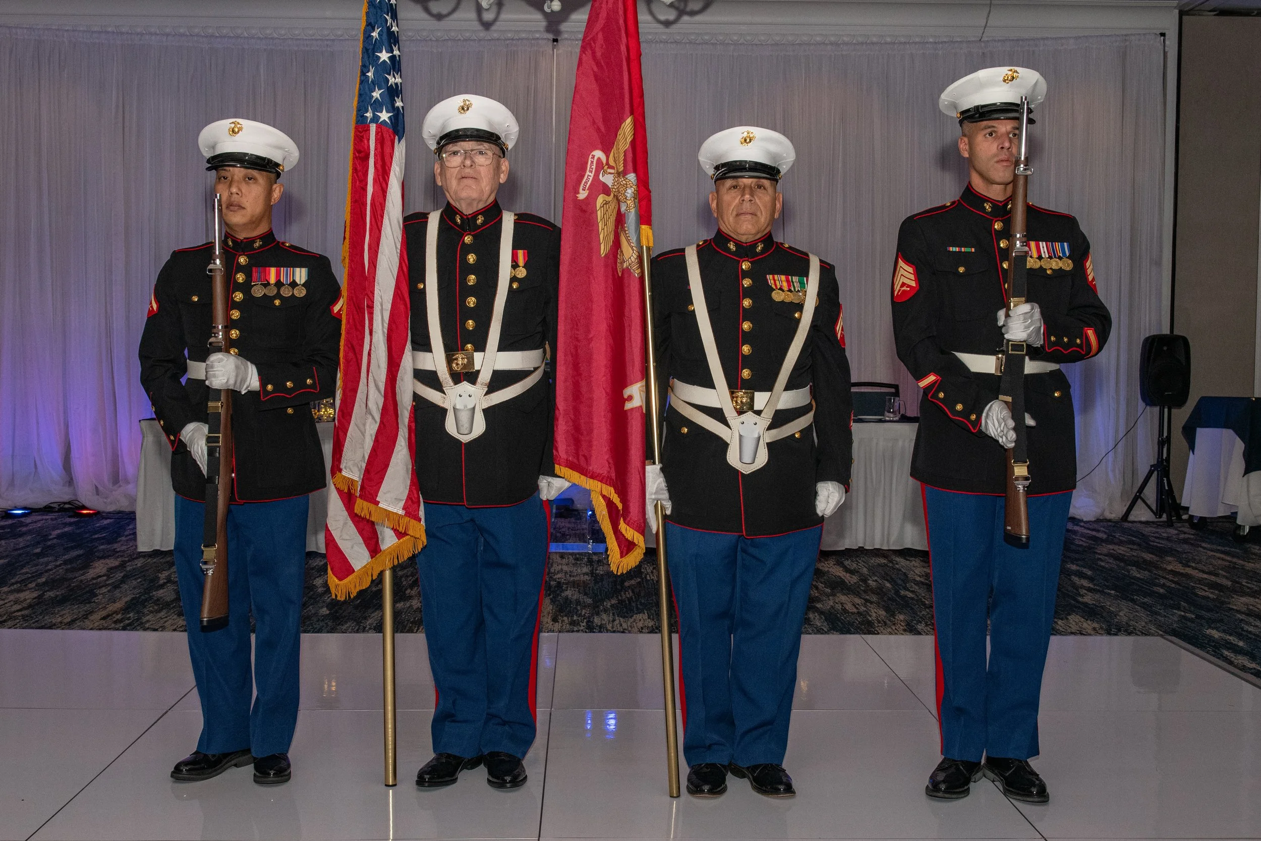 Four military personnel in dress uniform standing in a line indoors. The two individuals in the middle are holding flags, and the two on the ends are holding rifles. All are wearing white hats, black jackets with medals, and blue trousers with red st