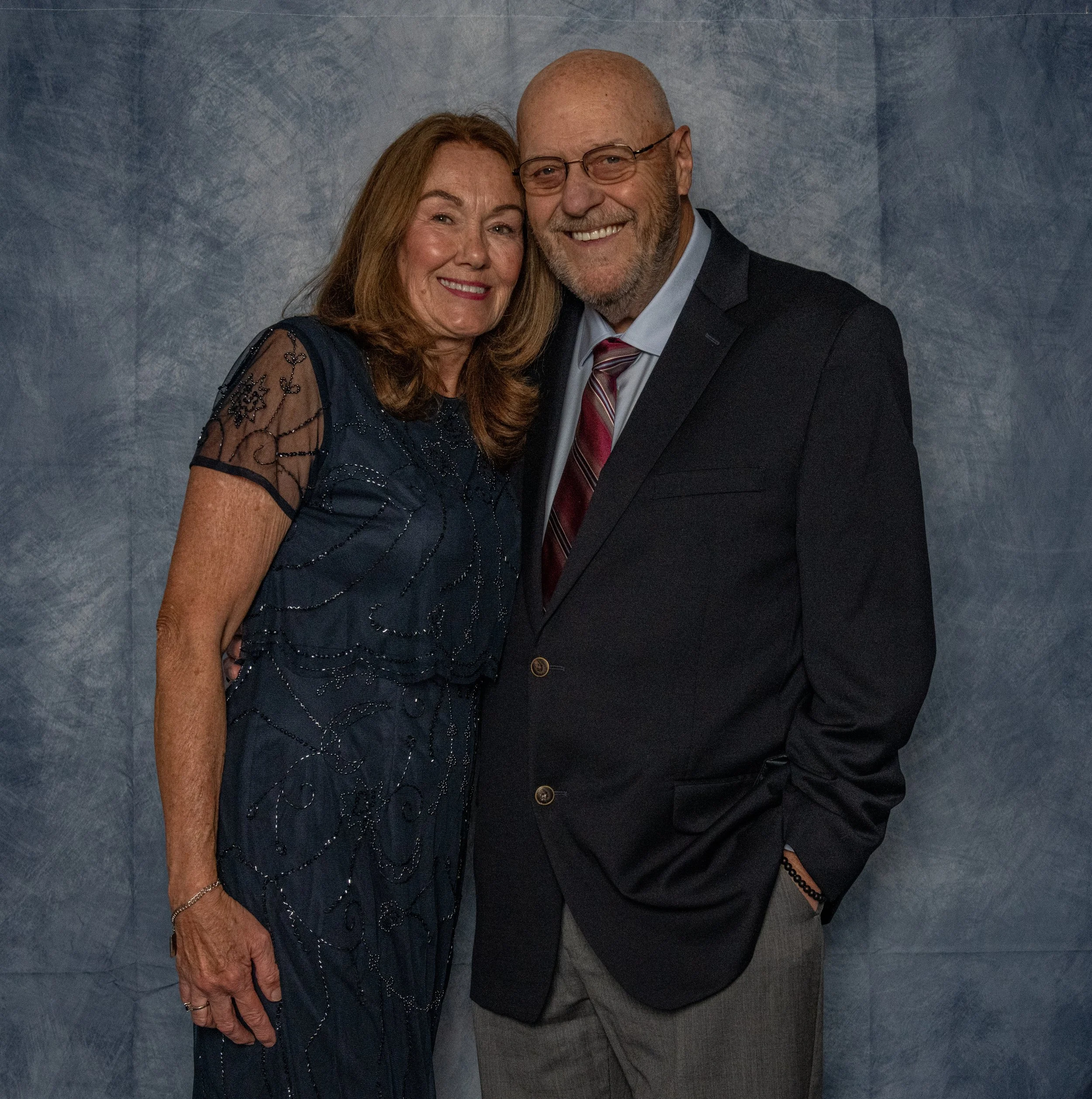 A smiling middle-aged woman in a navy blue dress with sheer and embroidered details, and a smiling middle-aged man in a dark suit with a pink striped tie, standing close together against a blue textured background.