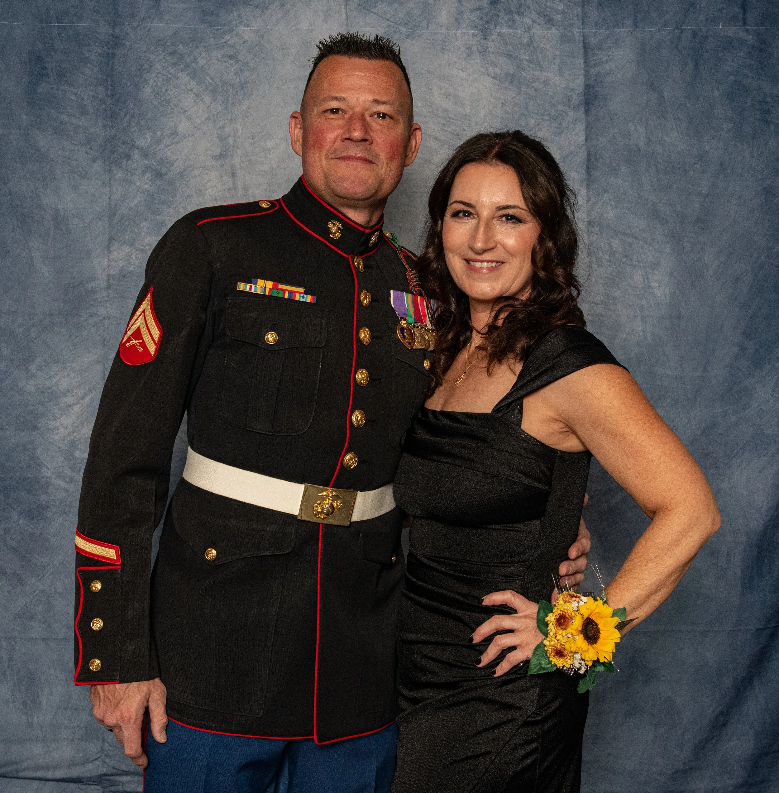A man in a United States Marine Corps dress uniform standing next to a woman in a black dress with a sunflower corsage, both smiling in front of a blue textured backdrop.
